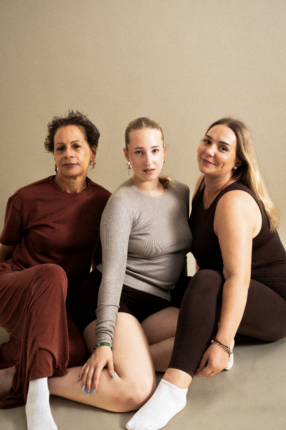 Trois femmes assises ensemble sur un sol neutre, regardant vers l'appareil, avec un mur beige en arrière-plan.