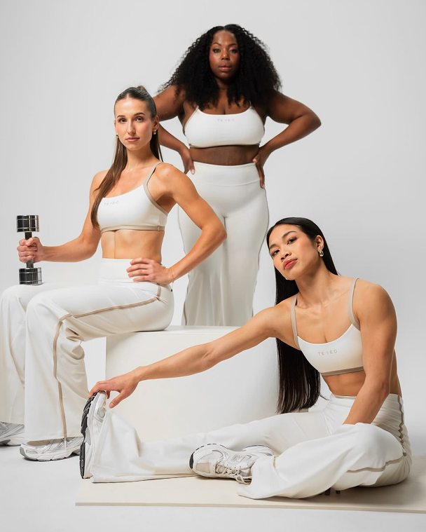 Trois femmes en vêtements de sport blancs posent devant un fond blanc. L'une d'elles tient un haltère, une autre s'étire en touchant sa chaussure, la troisième se tient confiée avec les mains sur les hanches.