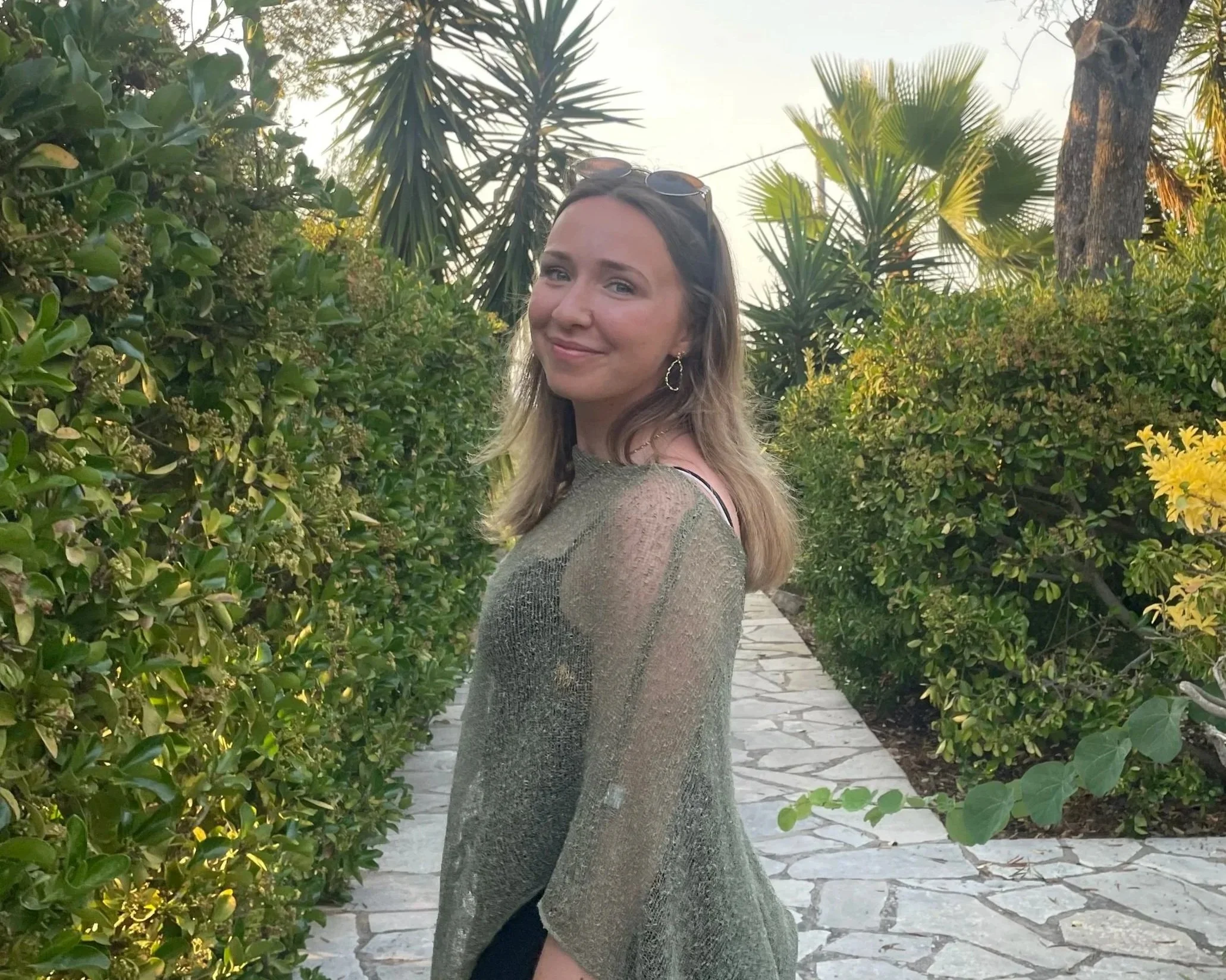 A young woman with long blonde hair and sunglasses on her head smiling and standing between green bushes and trees on a stone pathway.