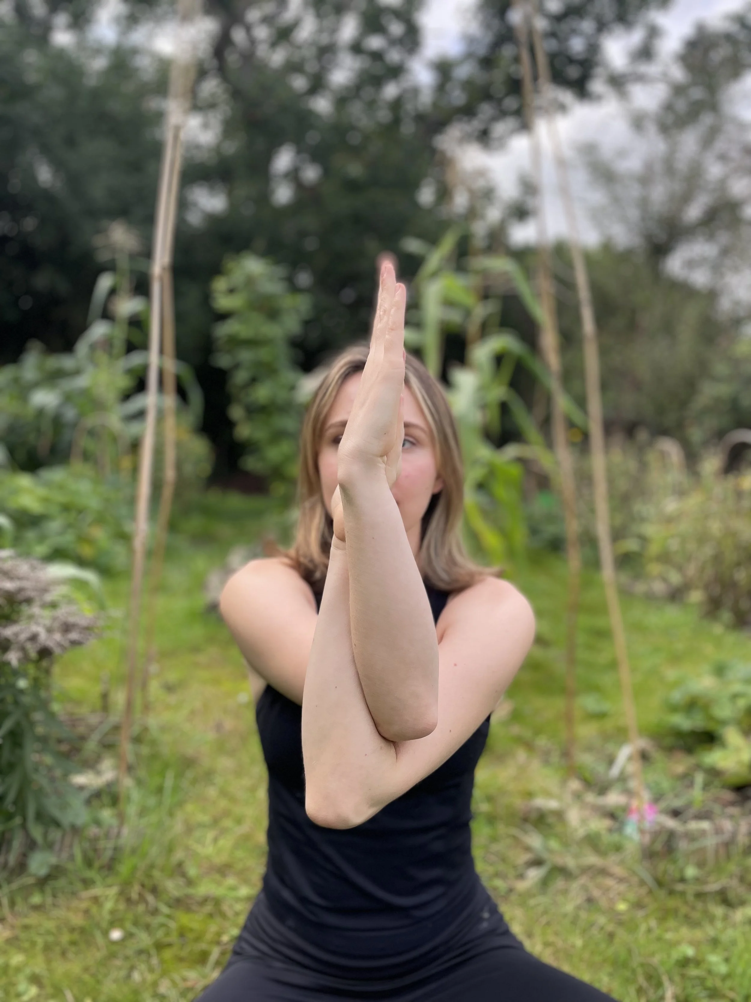 A woman practicing yoga outdoors, performing a pose with her arms crossed in front of her face, surrounded by greenery and plants.