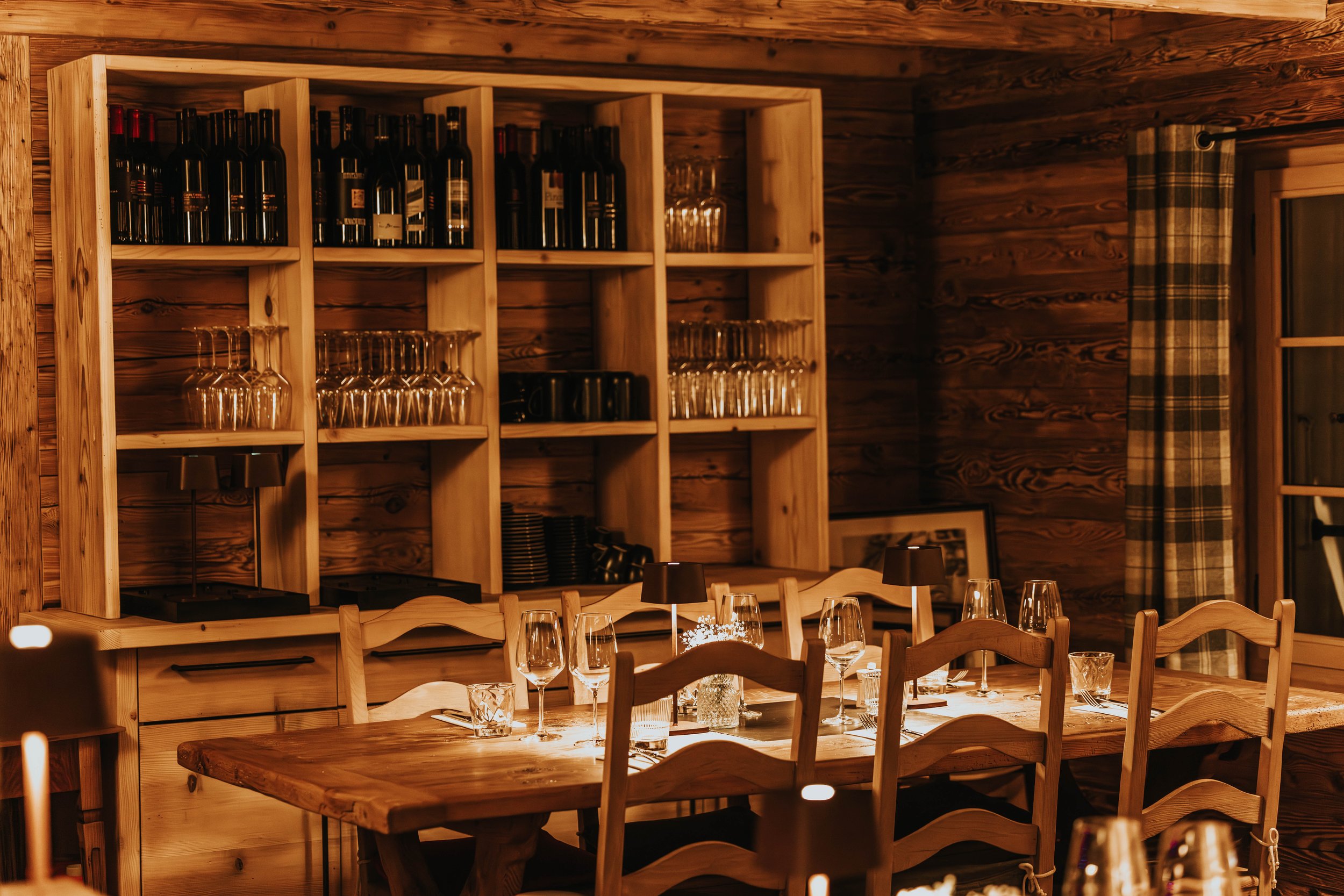 Intérieur d'une salle à manger avec murs en bois, une étagère avec des bouteilles et des verres, tables prêtées pour un repas avec des verres à vin et des couverts.