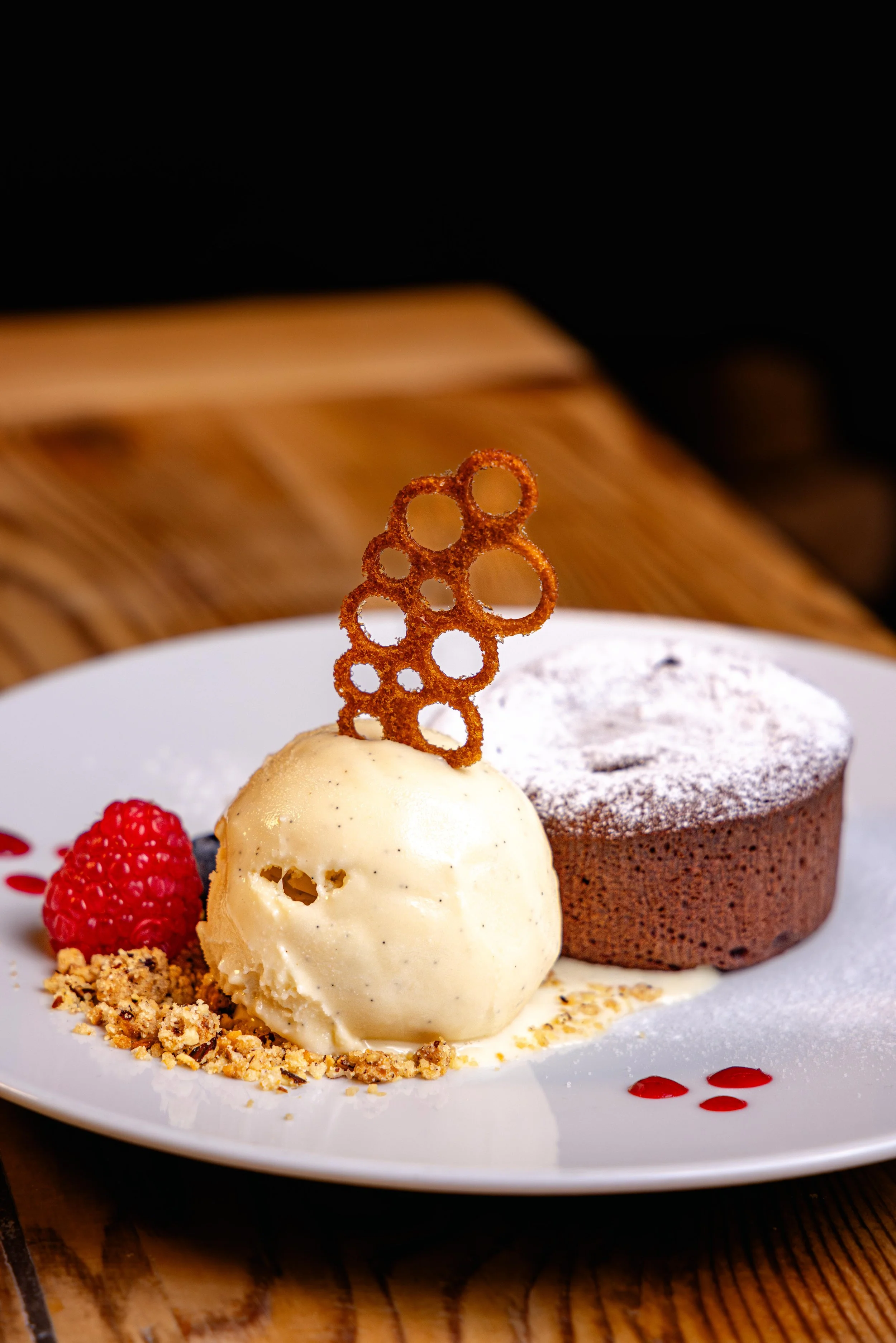 Un dessert avec une boule de glace vanille, un gâteau au chocolat, des framboises, des éclats de noisettes, et un décor de caramel en forme de cercle, présenté sur une assiette blanche.