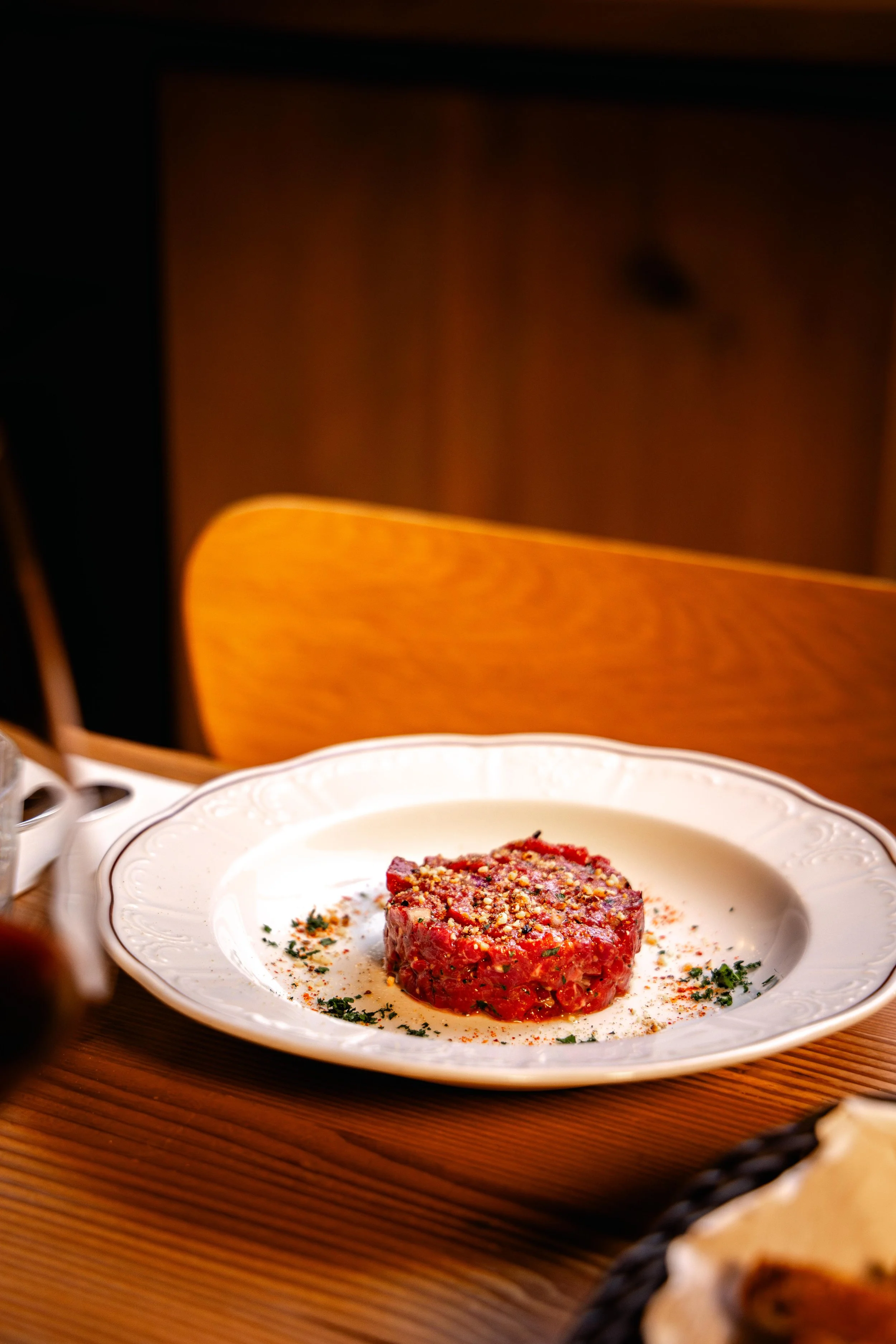 Un plat de viande hachée ou un steak tartare assaisonné sur une assiette en porcelaine blanche. En arrière-plan, une table en bois et une chaise en bois avec dossier rembourré.