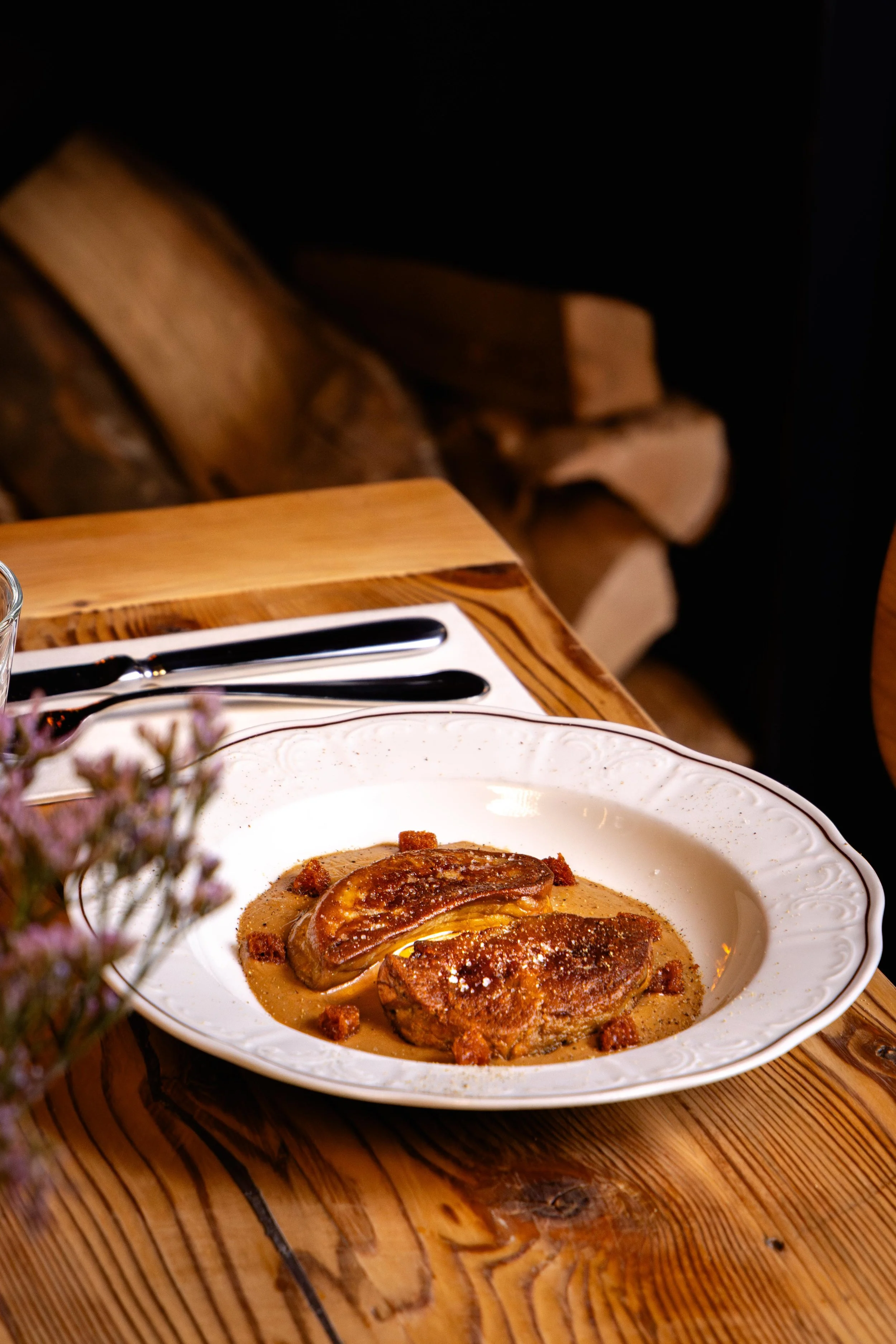 Une assiette blanche avec un plat de viande en sauce, accompagnée de morceaux de viande ou de garniture, sur une table en bois. En arrière-plan, un lot de bois empilé et une nappe blanche avec un couteau et une fourchette posés dessus.