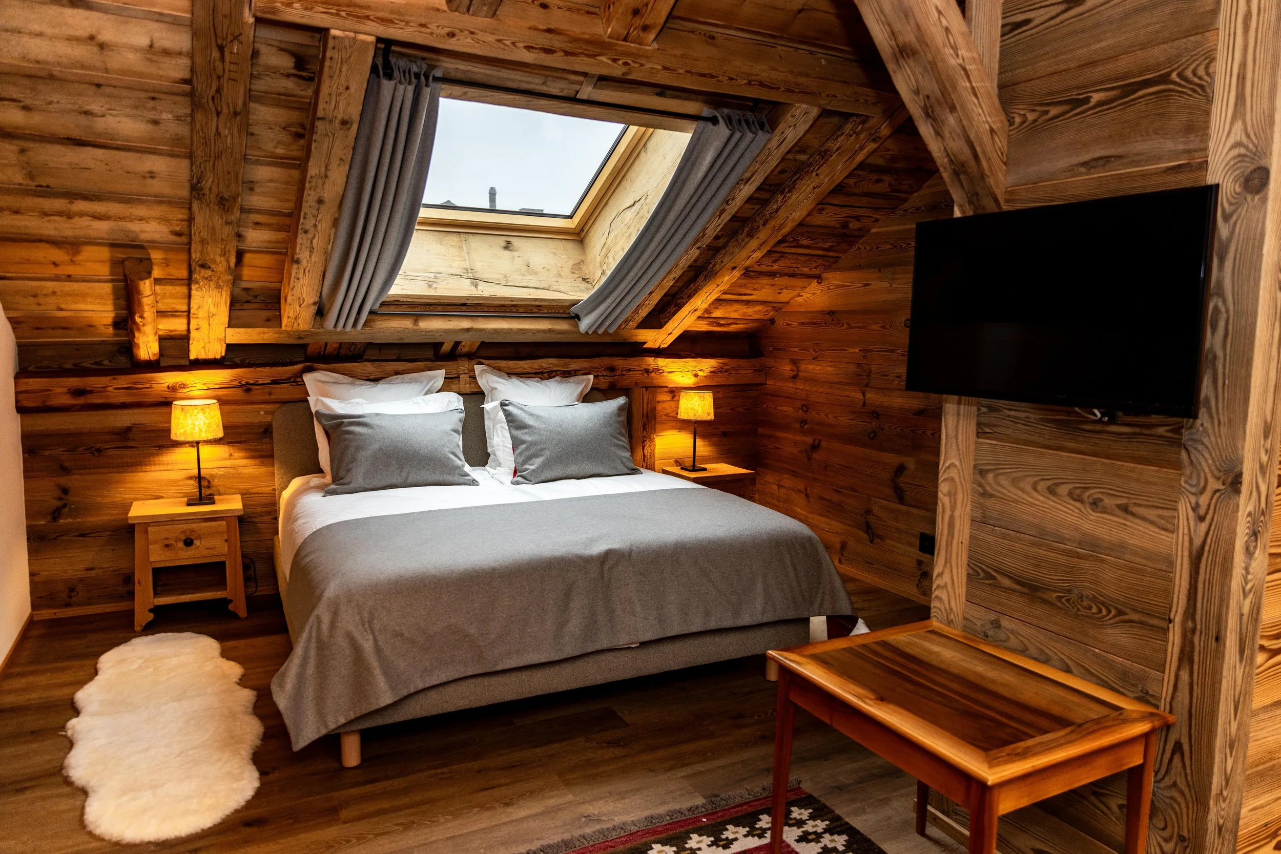 Chambre avec murs et plafond en bois, lit avec draps gris et coussins blancs et gris, deux lampes de chevet allumées, tapis en peau de mouton blanc, fenêtre de toit laissant entrer la lumière naturelle, téléviseur sur le côté droit.