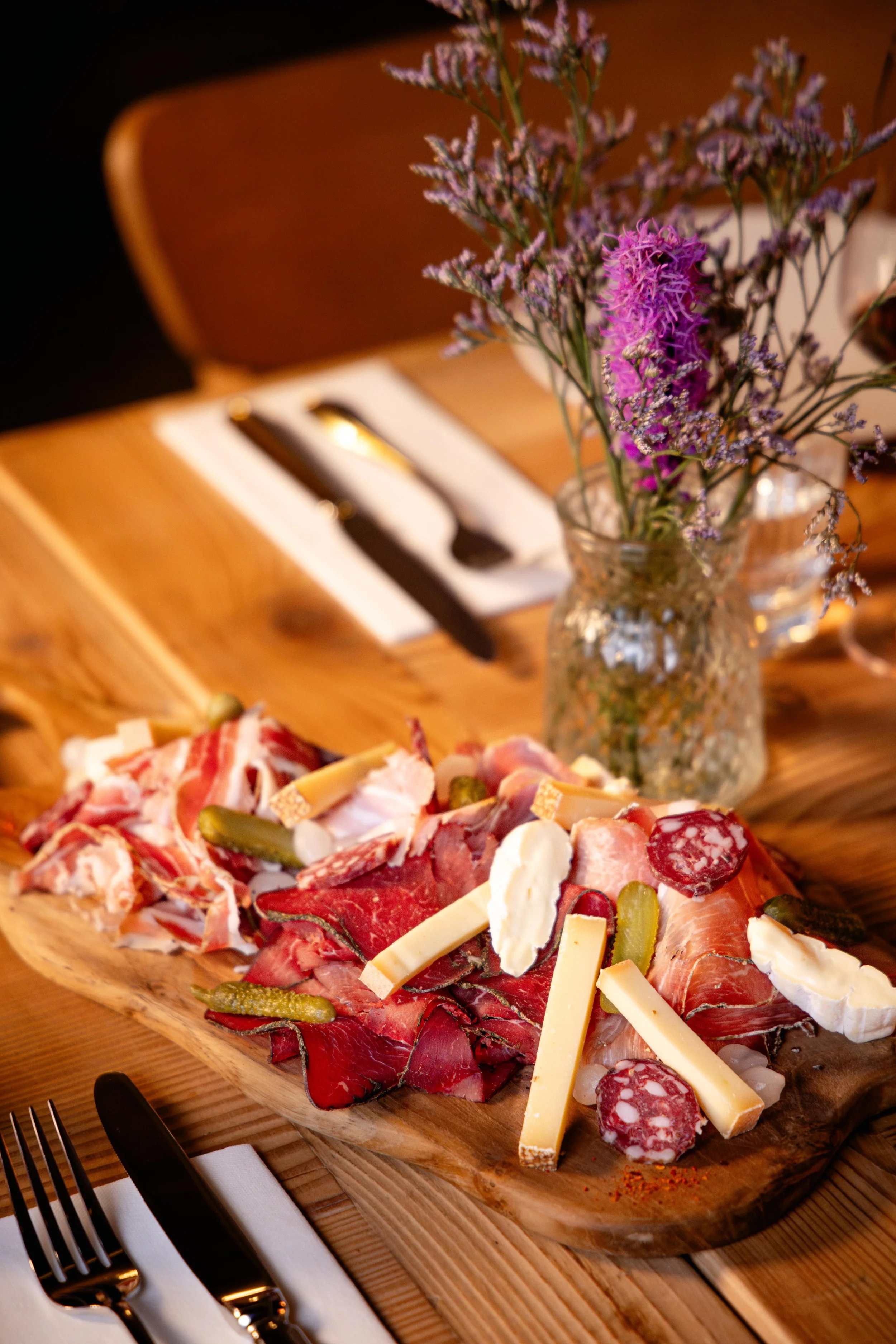 Planche de charcuterie avec différentes viandes, fromages, cornichons, sur une planche en bois, avec un vase de fleurs violettes et roses, et des ustensiles en arrière-plan.