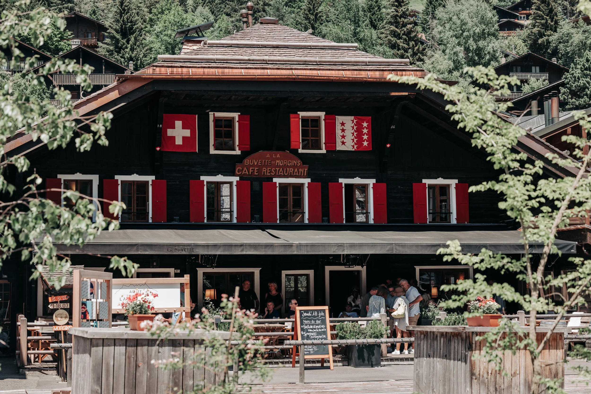 Une auberge en bois avec des drapeaux suisses et chatterie, des personnes assises à une terrasse extérieure, entourée de verdure montagnarde.