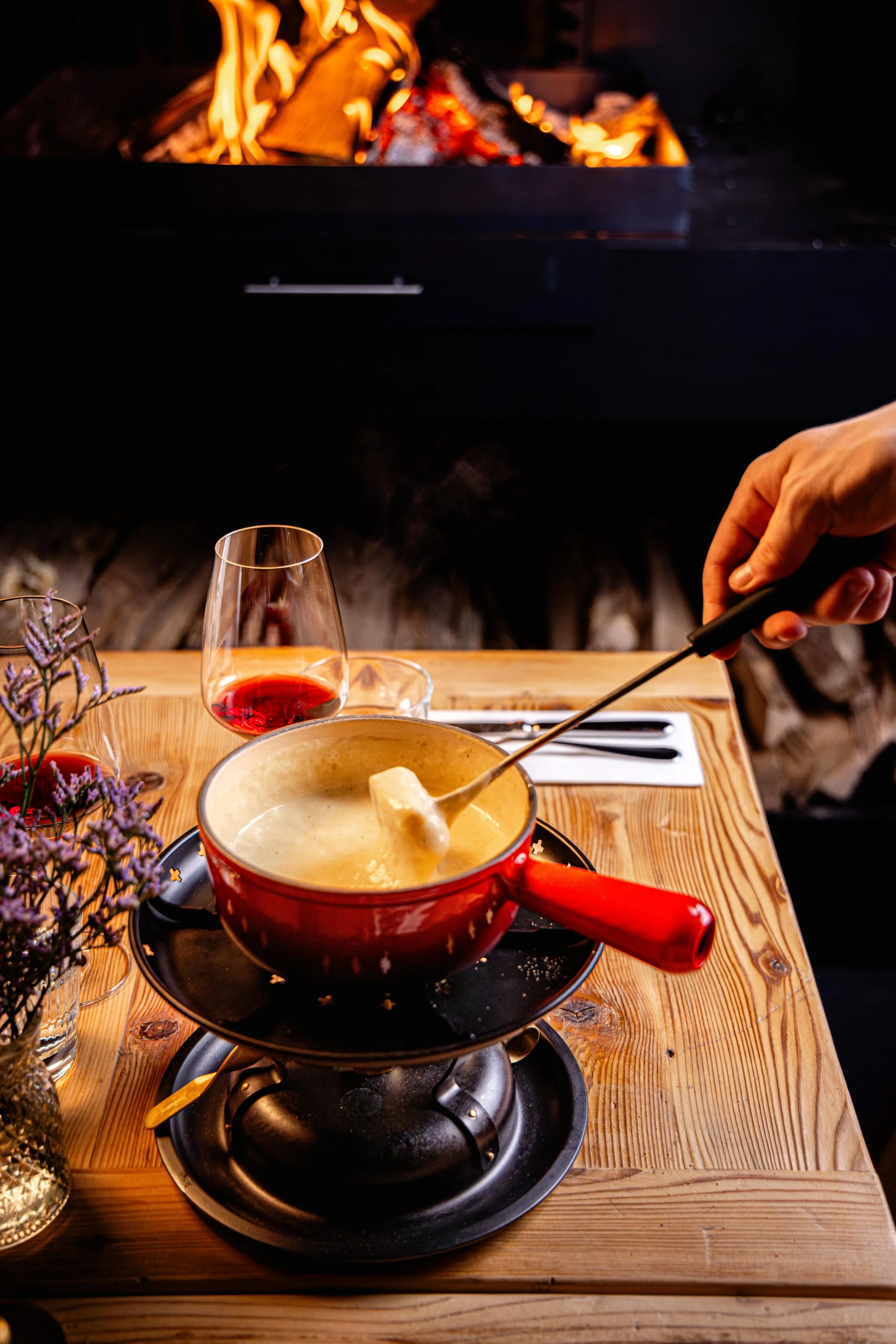 Une fondue au fromage dans une casserole rouge sur un réchaud, avec un verre de vin rouge à côté, sur une table en bois, avec des couverts et des fleurs violettes.