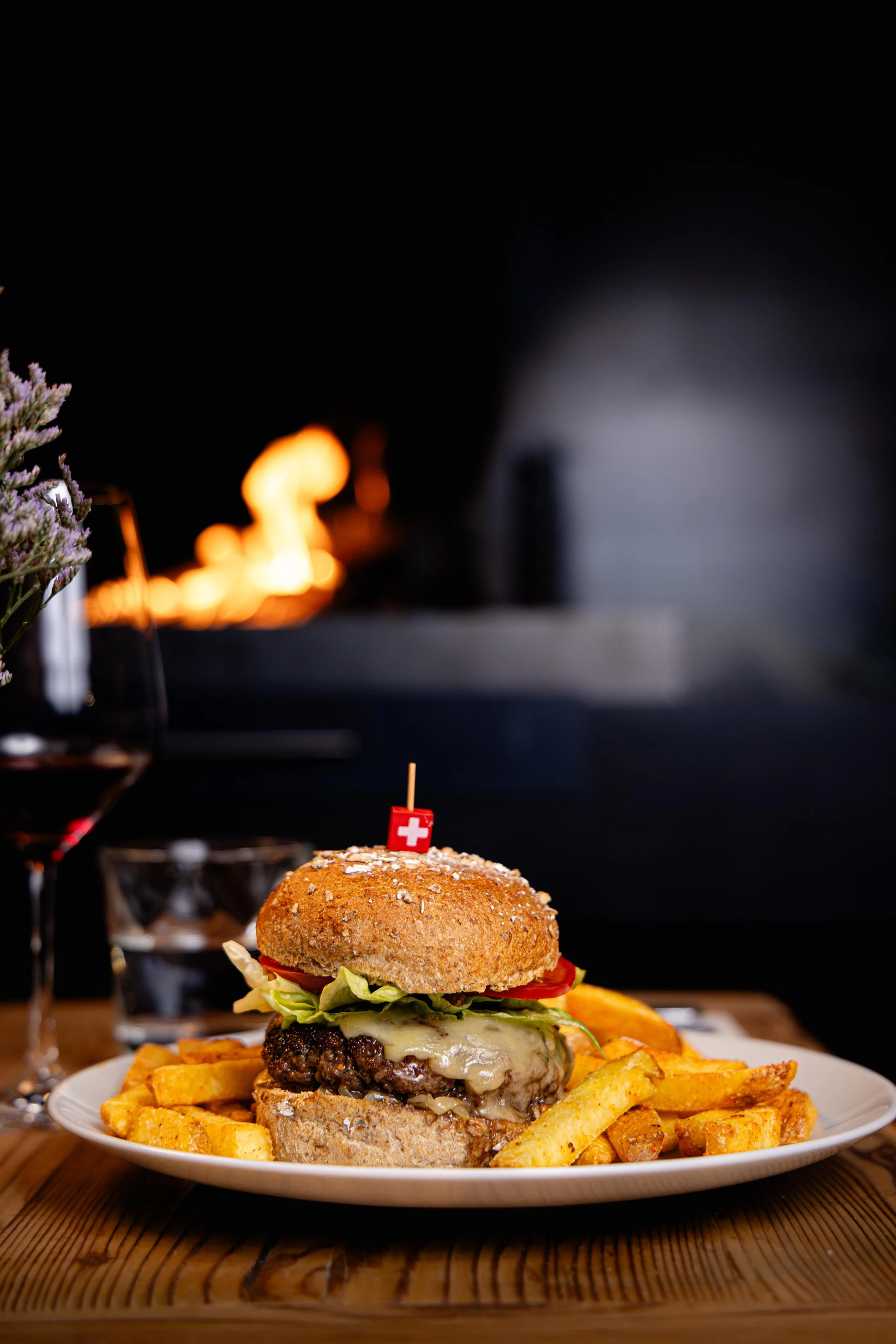 Un cheeseburger avec des frites sur une assiette, avec un drapeau suisse inséré dans le burger, placé sur une table en bois, avec un fond sombre et une flamme de feu en arrière-plan.