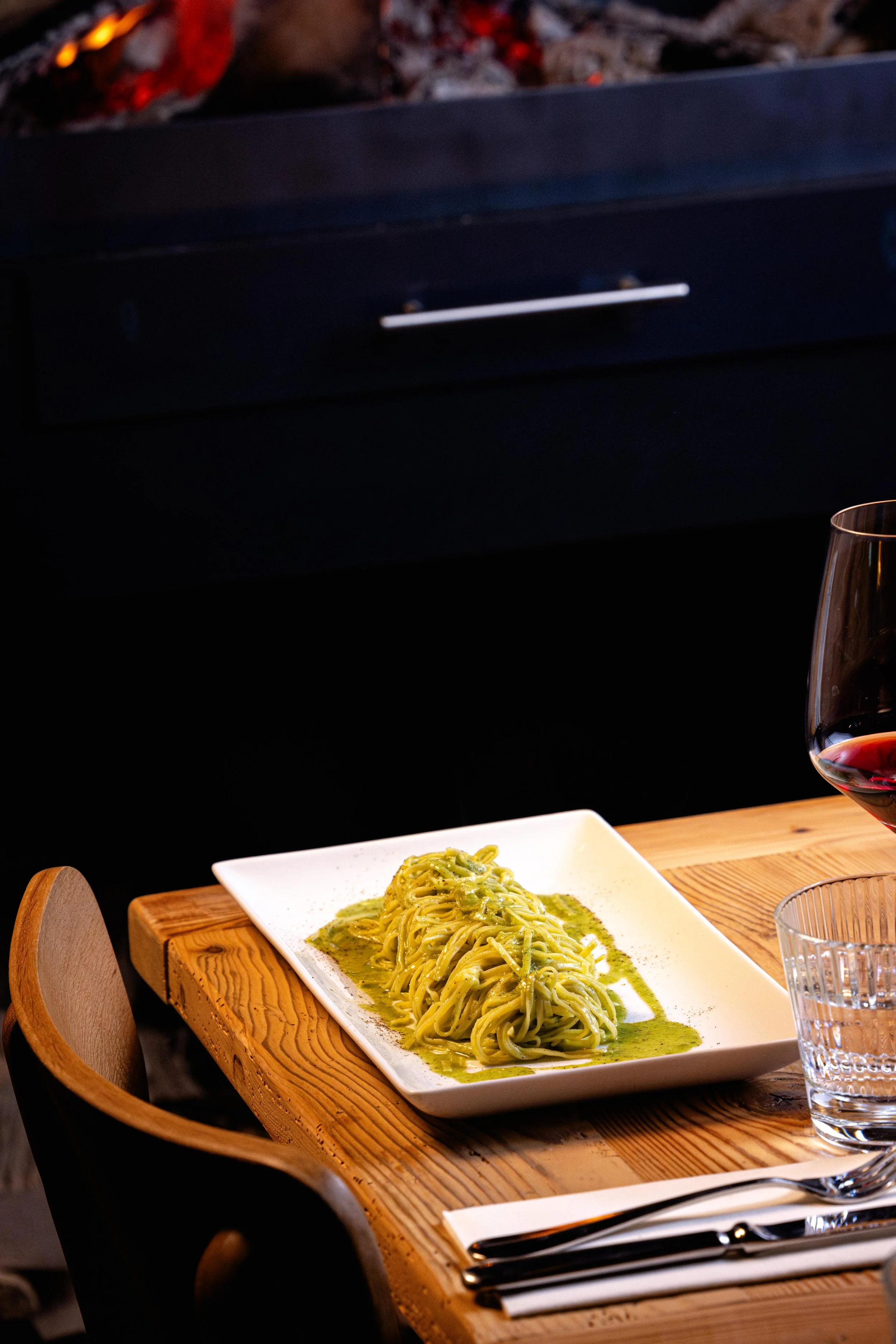 Plat de pâtes vertes avec sauce pesto, un verre de vin rouge, et un verre d'eau, à une table en bois dans un restaurant élégant.