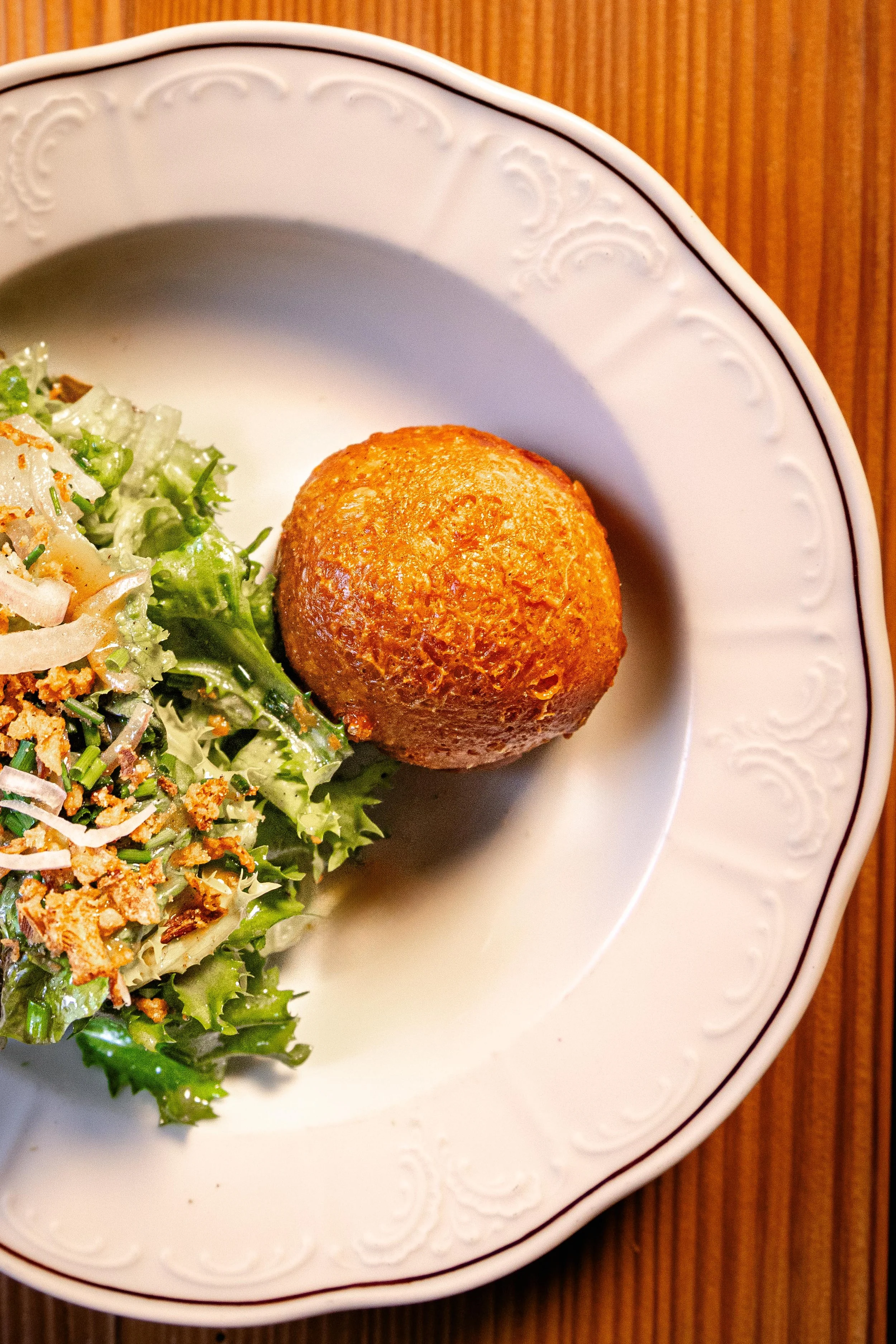 Une assiette blanche avec une bordure décorative, contenant une salade verte et une boule de fromage croustillante, posée sur une surface en bois.
