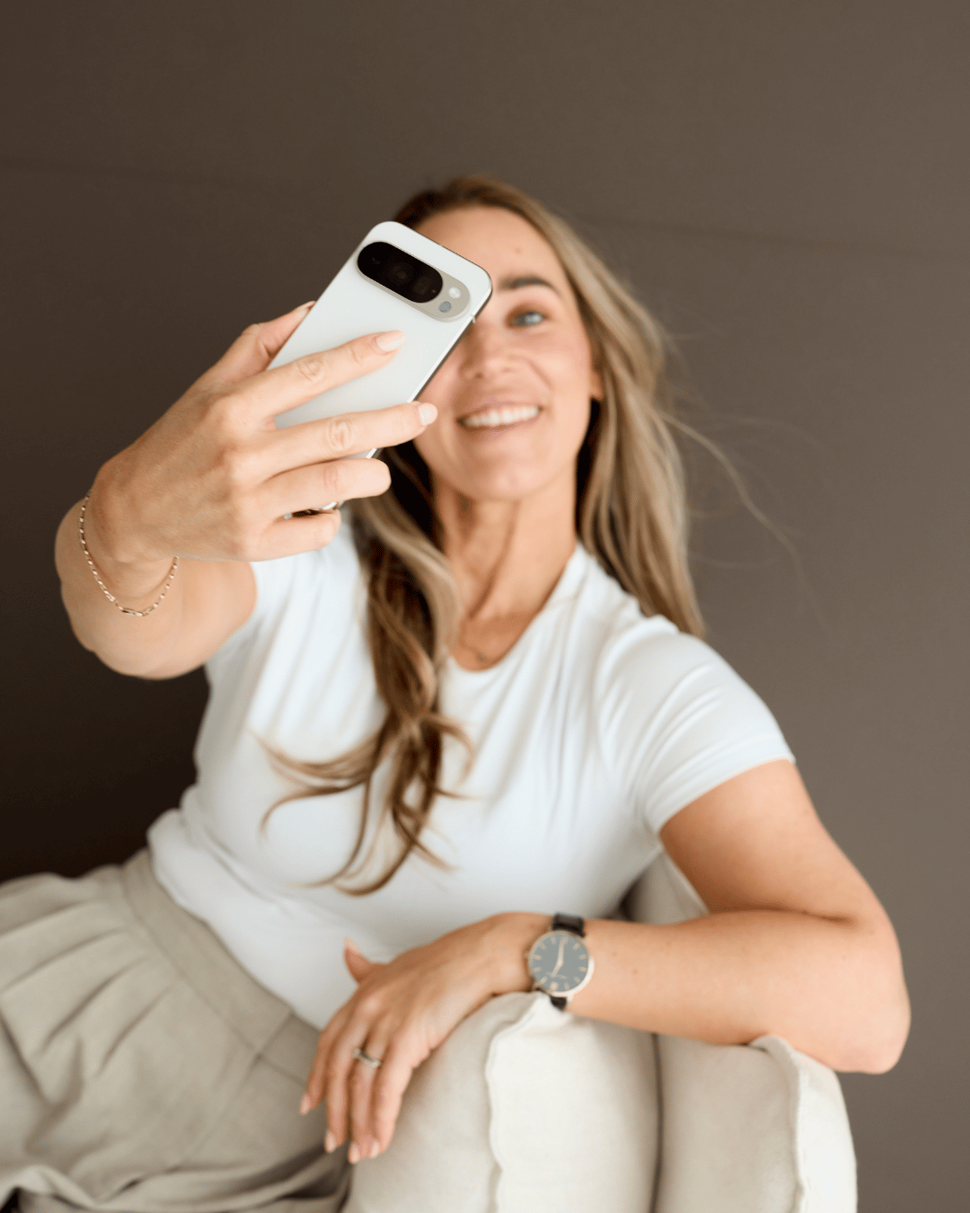 A woman taking a selfie with a smartphone, smiling, sitting on a beige armchair, wearing a white t-shirt and beige pants, with a black watch on her left wrist and a silver bracelet on her right wrist.