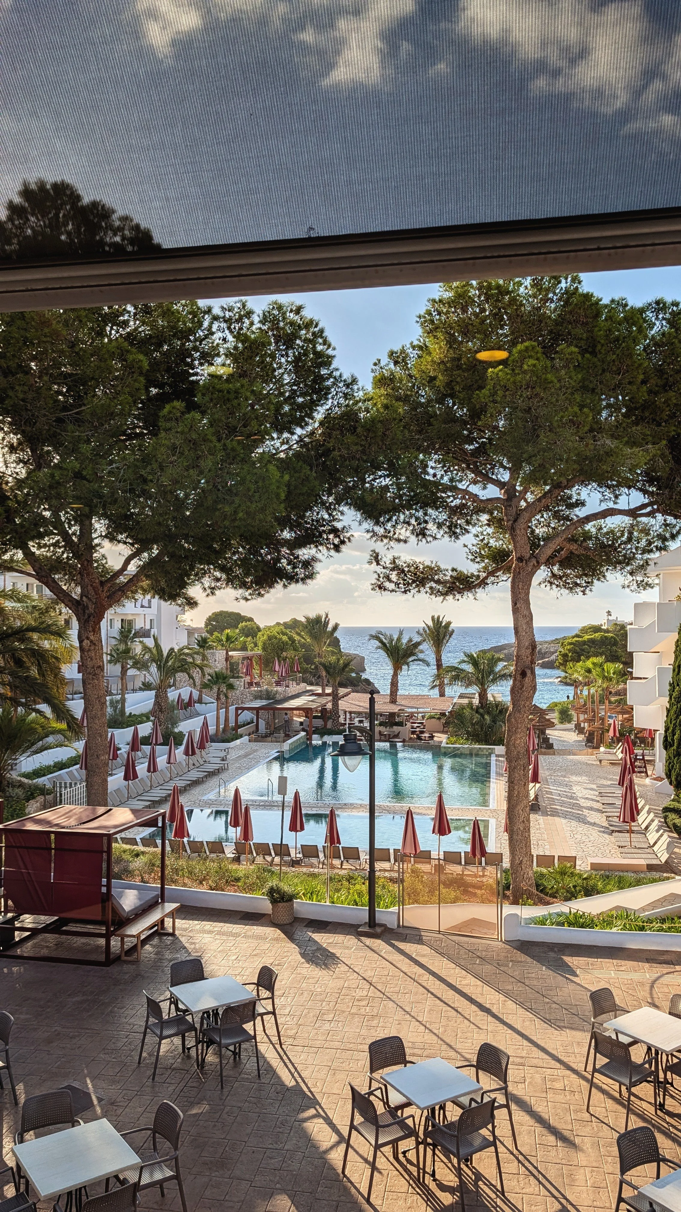 View of a resort pool area with red umbrellas, lounge chairs, greenery, trees, and the ocean in the background, seen from a shaded balcony with tables and chairs.