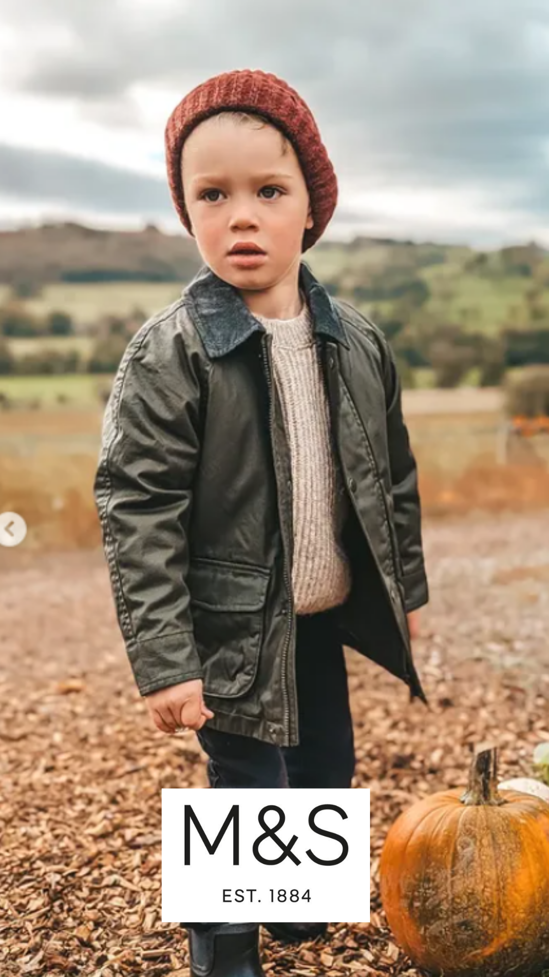 A young boy standing outdoors on a fall day, wearing a red knit hat, a gray leather jacket, and a beige sweater, with a pumpkin on the ground nearby, and an M&S logo on the bottom of the image.