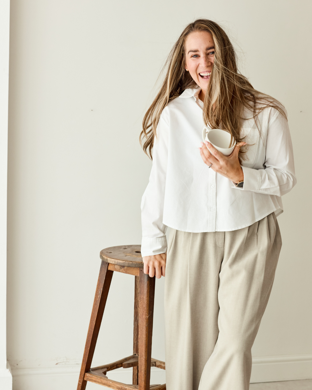 A woman with long, light brown hair smiling and holding a white mug, standing next to a wooden stool in a room with beige walls.