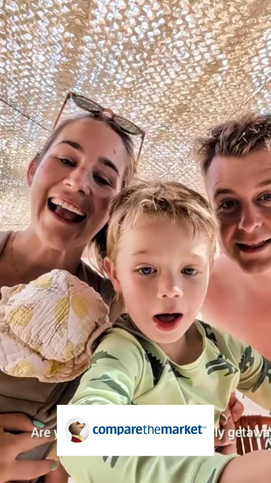 A group of three people, two adults and one child, taking a selfie underneath a decorative lattice ceiling. The two adults are smiling and the child looks surprised or excited. The woman has sunglasses on her head and is holding the child, the man is on the right side of the image.