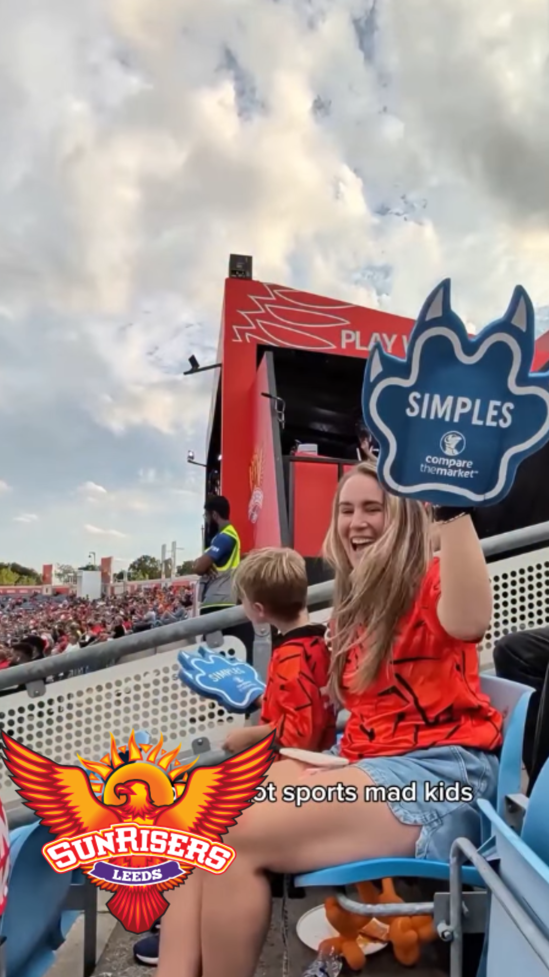 A woman at a sports game, smiling and holding a blue foam finger labeled 'SIMPLES'. She has long blonde hair and is wearing a red shirt. Next to her is a young boy in a red and black shirt, sitting on a bench. The crowd is visible in the background, and the sky is partly cloudy. The logo of the Sunrisers Leeds cricket team is overlaid on the bottom left of the image.