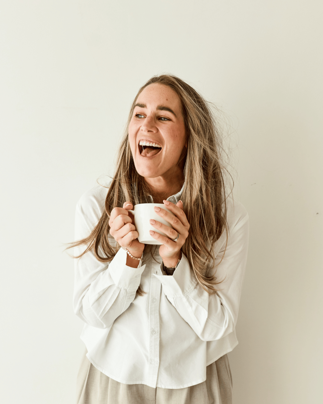 Woman with long hair smiling and laughing while holding a white mug, wearing a white shirt, standing against a plain light-colored background.