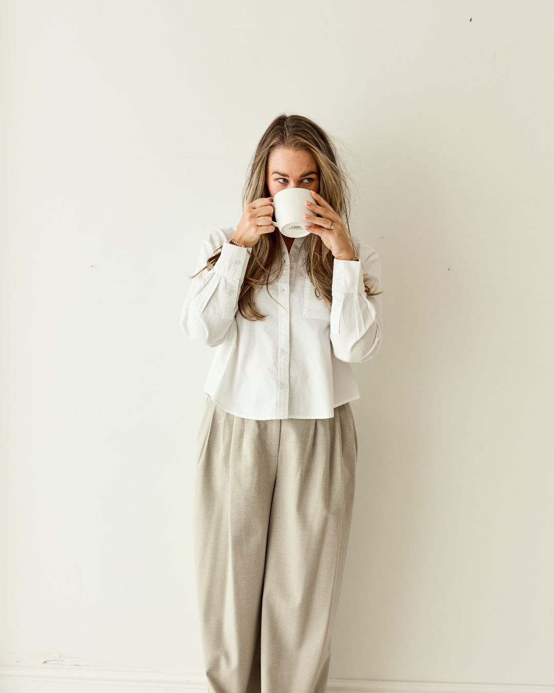 Woman with long blonde hair drinking from a white mug, standing against a plain white wall.