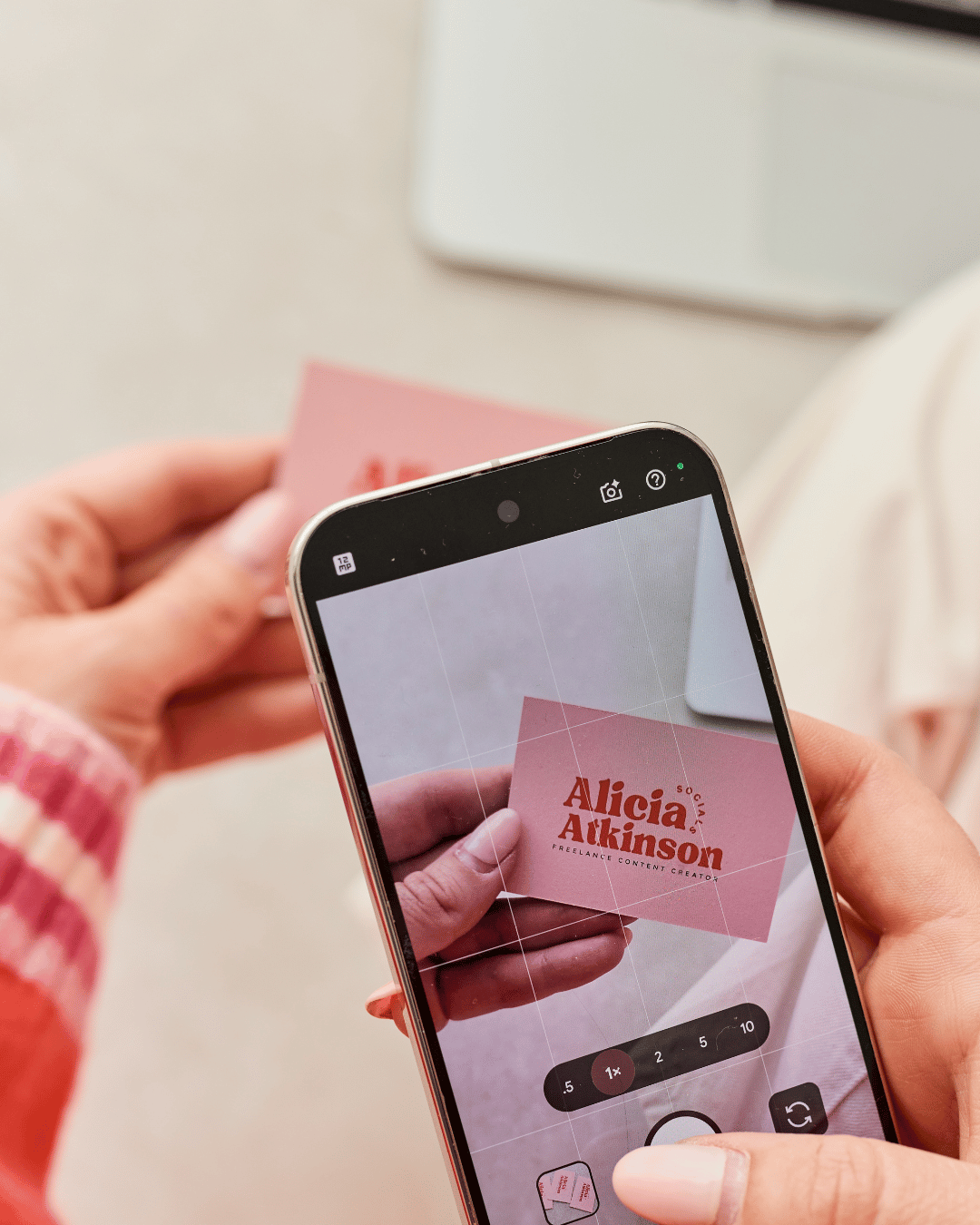 Person taking a photo of a pink business card with the name 'Alicia Atkinson' on her smartphone.