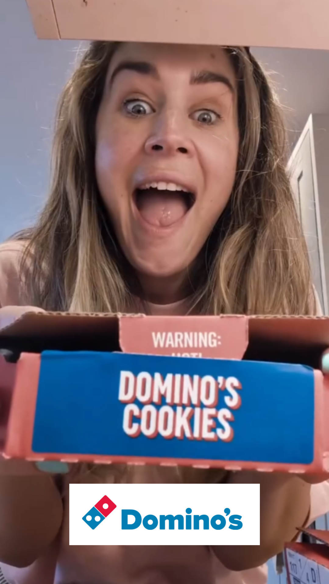 A woman with long hair looking down into a box of Domino's cookies, showing an excited expression.