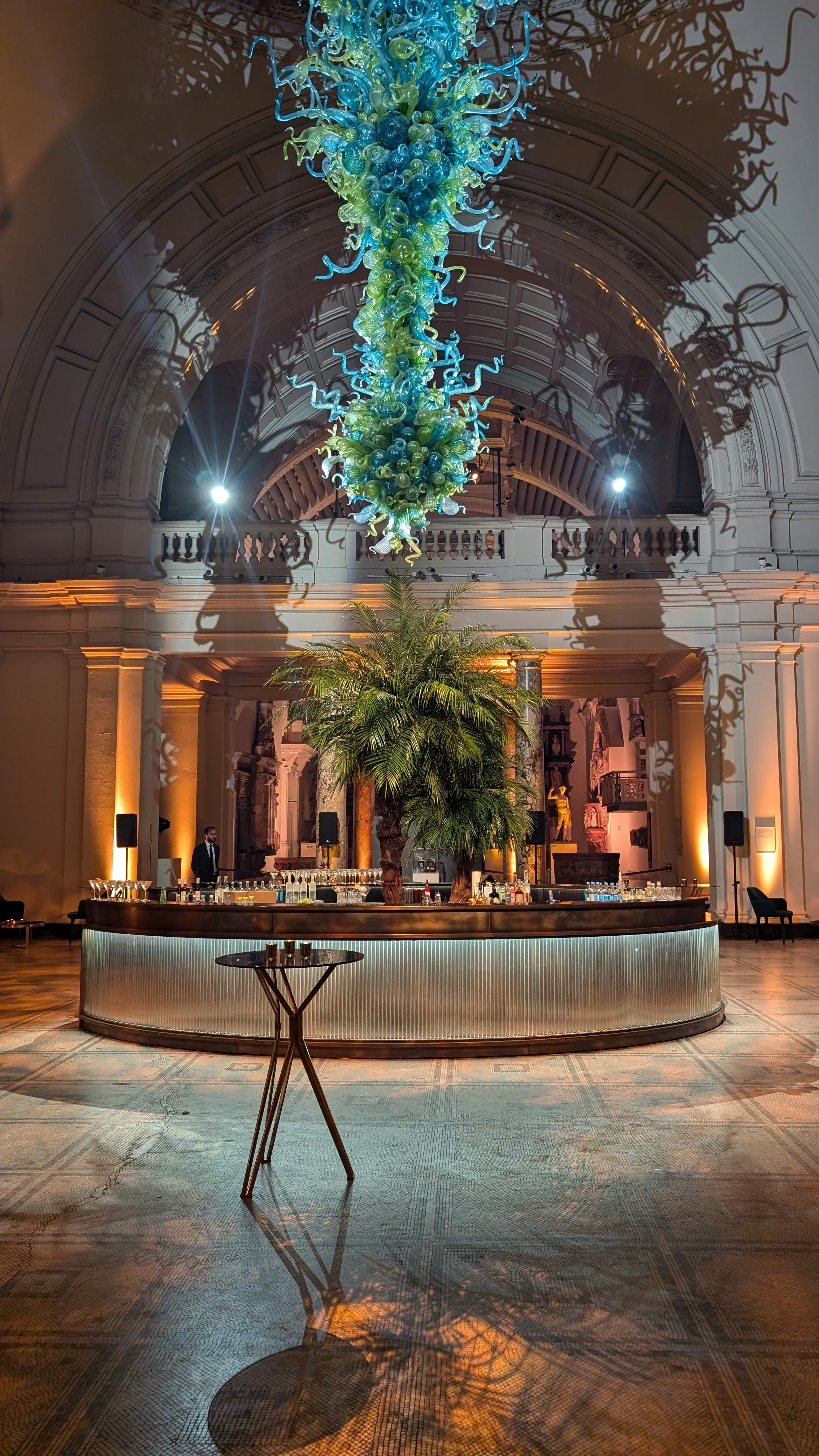Elegant indoor space with a large colorful chandelier hanging from the ceiling, casting intricate shadows. There is a bar area with a circular counter, a palm tree in the center, and warm lighting highlighting architectural features such as arches and decorative columns. Two staff members are standing behind the bar.