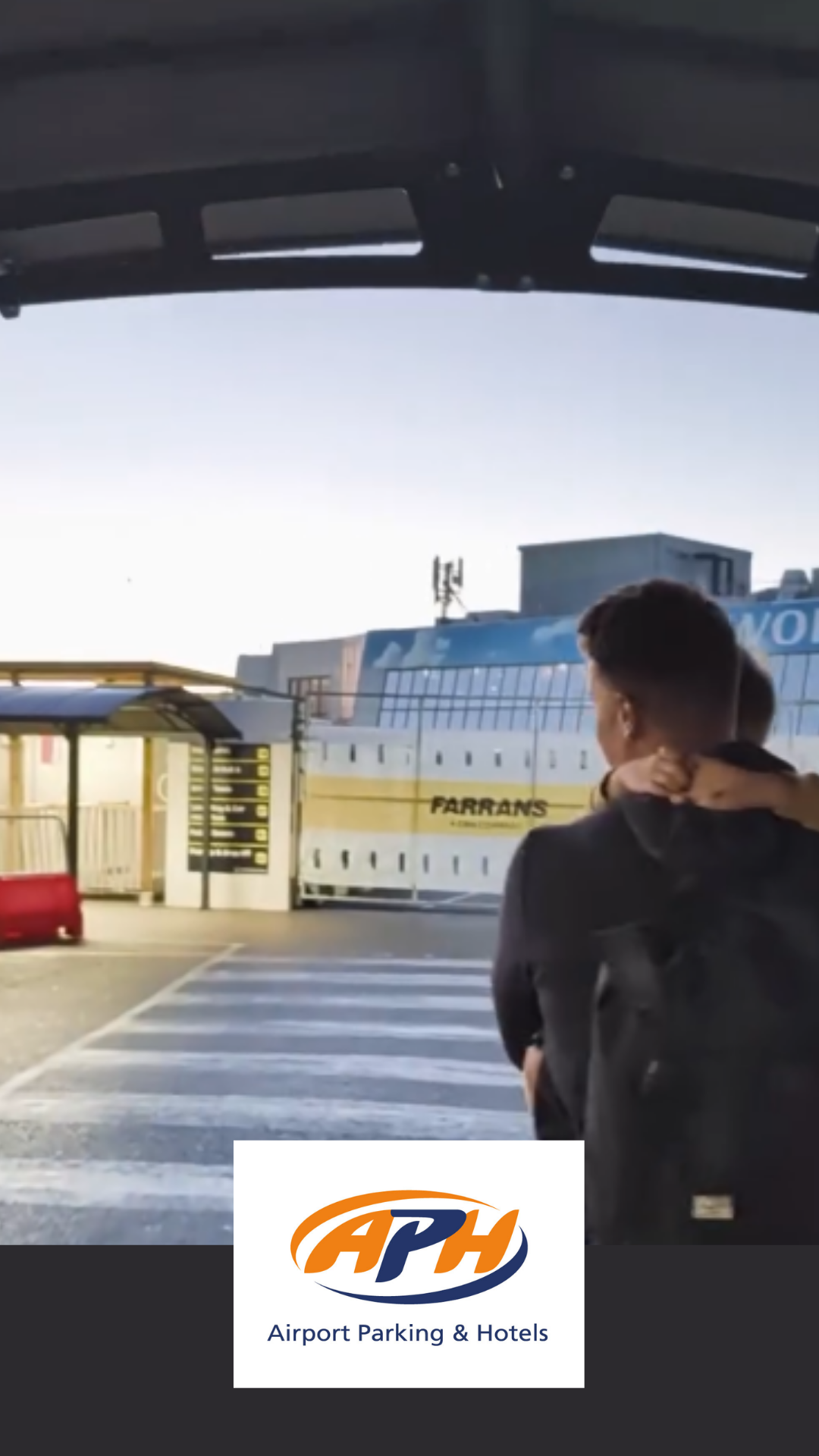 Two men hugging in an airport parking lot near a building with signs. Part of an airport shuttle bus and a crosswalk are visible. The logo of Airport Parking & Hotels is at the bottom of the image.