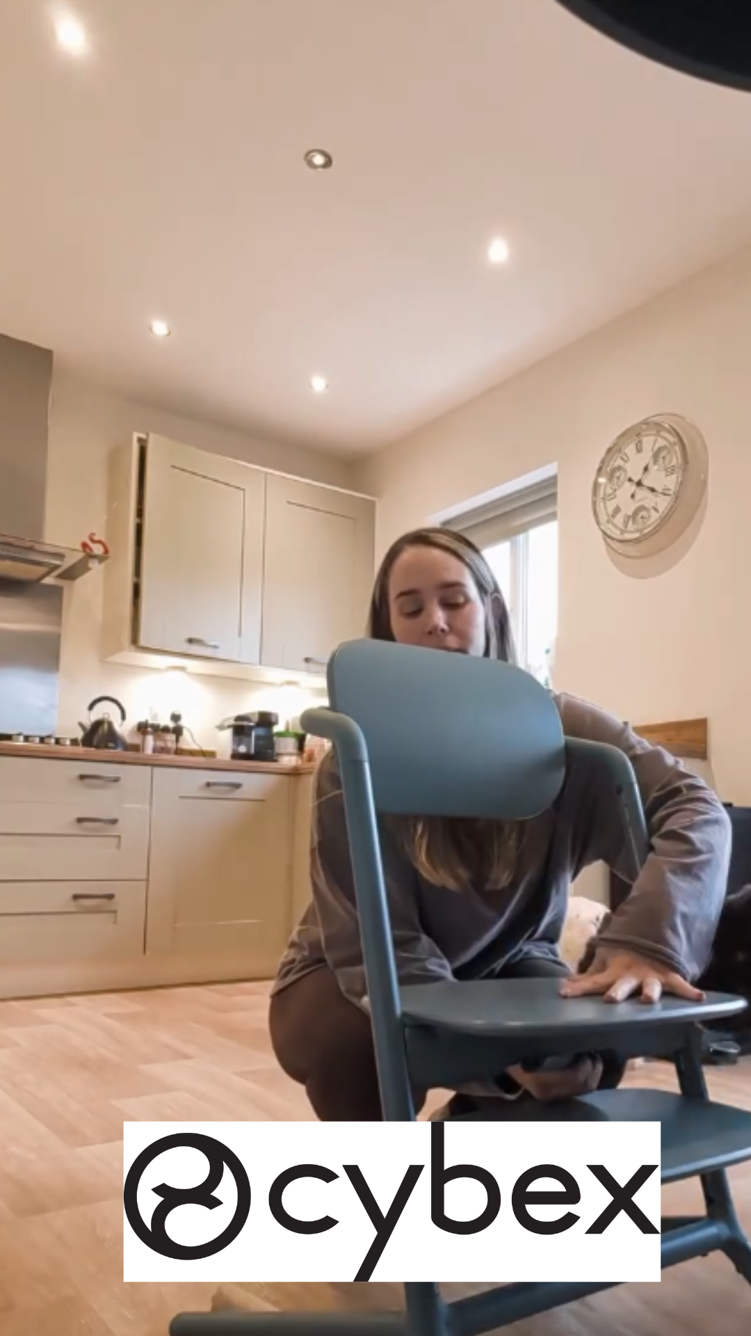 A woman in a kitchen kneeling on the floor, holding a green chair in front of her, with her mouth near the top of the chair backrest. The kitchen has cream-colored cabinets, a countertop with various kitchen items, a window, and a clock on the wall.