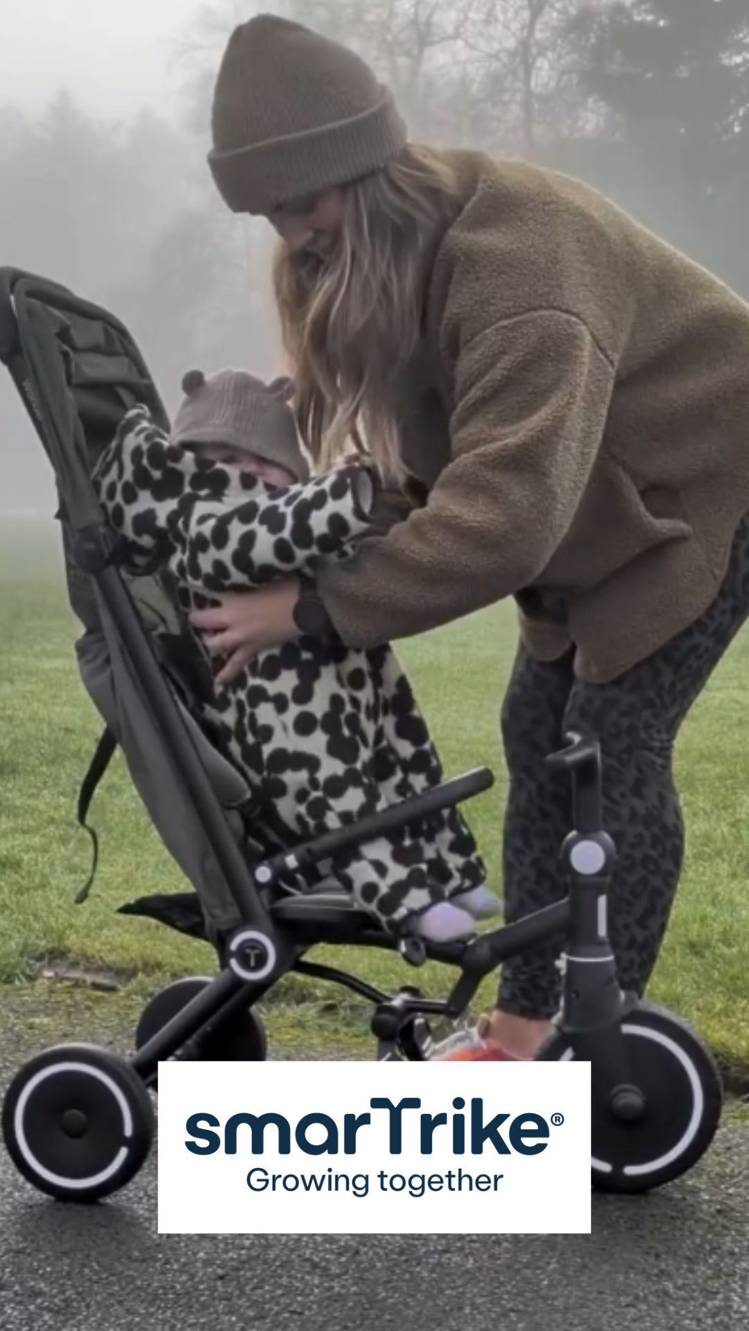 A woman in a beanie and cozy jacket helping a young girl in a animal print hoodie and matching beanie ride a black and gray tricycle outdoors on a foggy day.