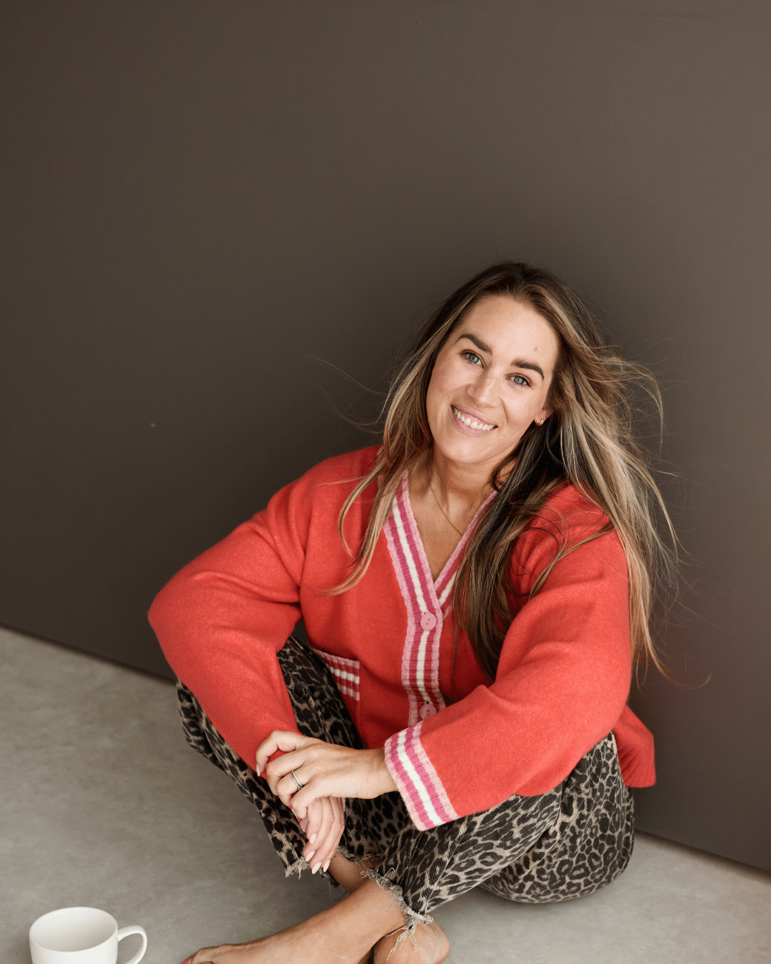 A woman with long, wavy hair sitting on the floor, smiling at the camera, wearing a red cardigan and animal print pants, with a white cup nearby, against a plain dark background.