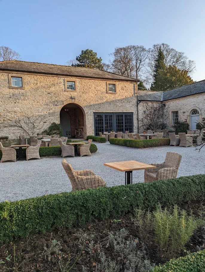 Outdoor patio with wicker chairs and tables, stone building with arched doorway and large windows, surrounded by neatly trimmed hedges and trees.