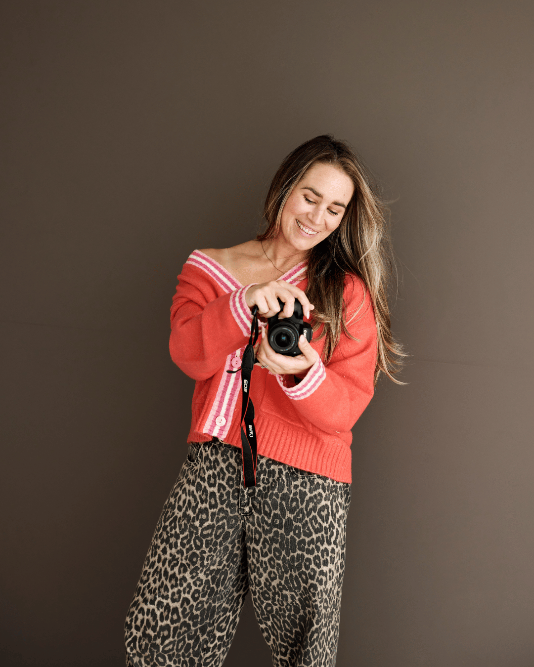 A smiling woman with long hair, wearing a red cardigan with pink and white stripes on the collar and cuffs, and leopard print pants, holding a camera and looking down at it against a plain dark background.