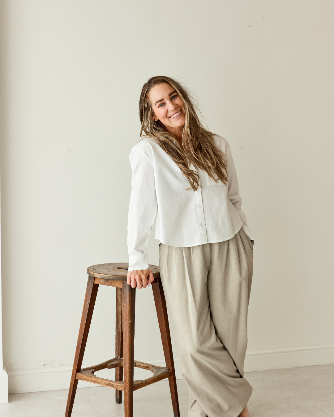 A young woman with long, wavy hair, smiling, wearing a white shirt and beige wide-leg pants, standing next to a wooden stool against a plain off-white wall.