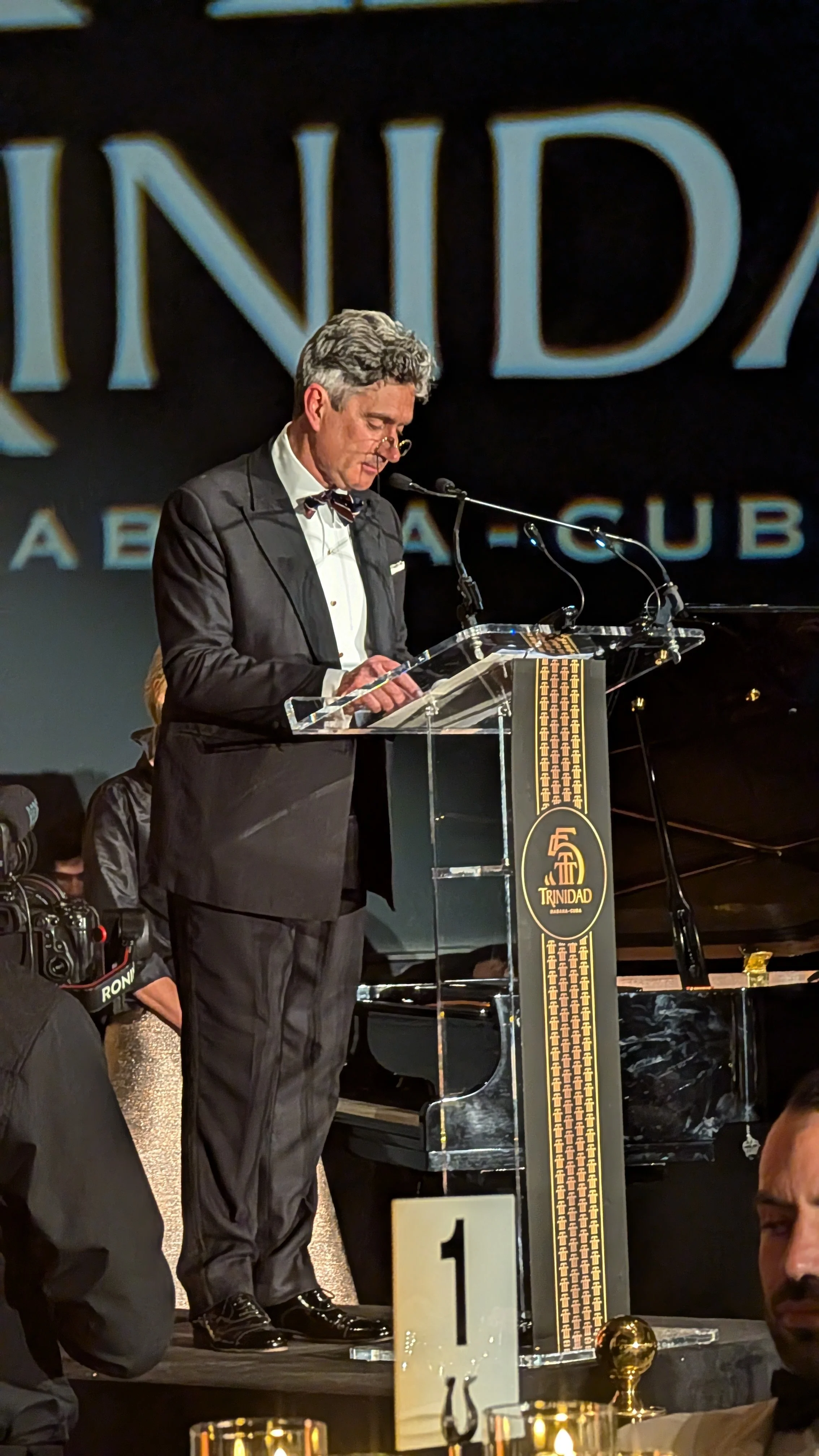 A man in a tuxedo with a bow tie and glasses stands at a clear podium with a microphone, reading or speaking during an event titled 'Trinidad' at a formal gathering or awards ceremony.