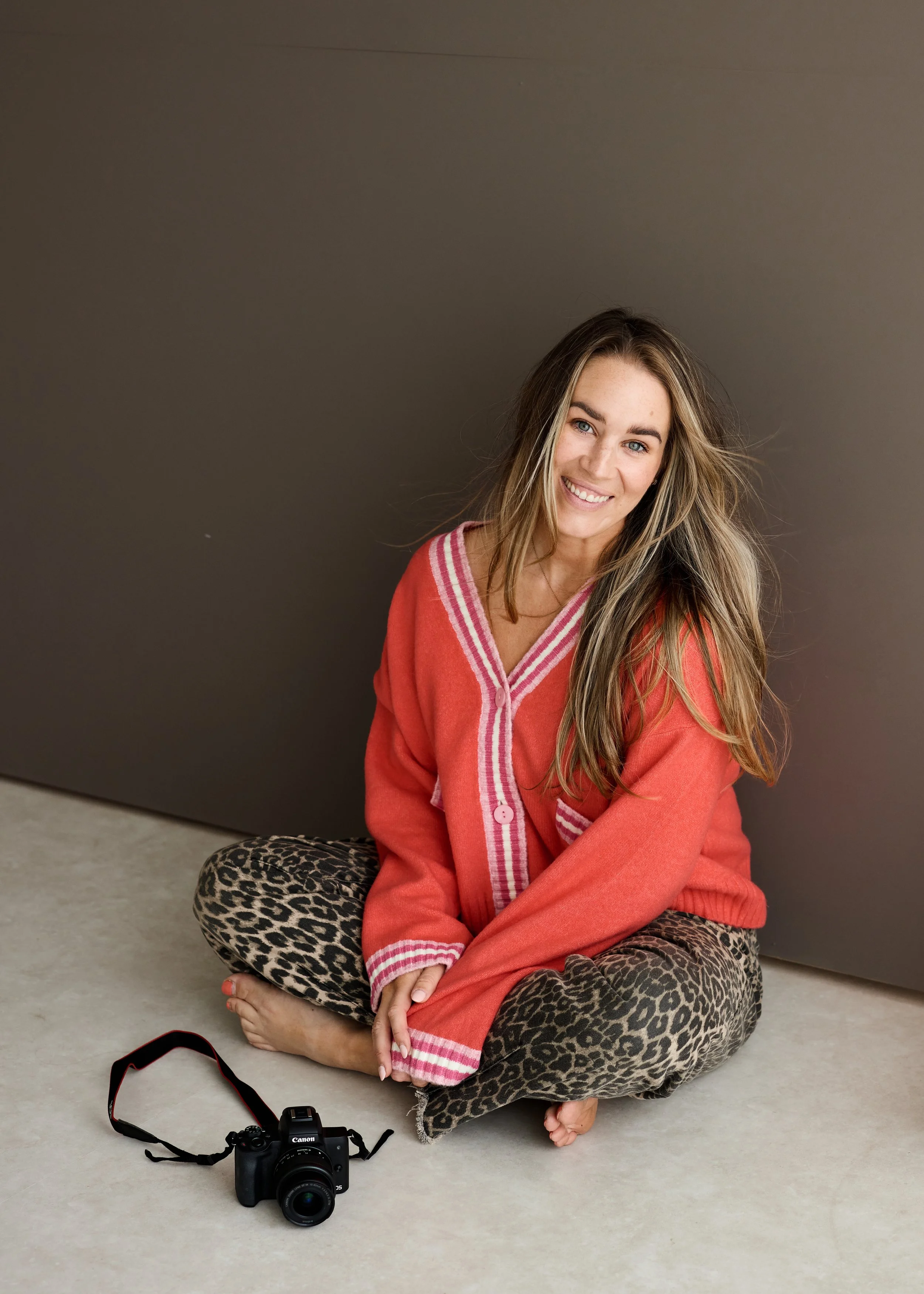 A woman with long blonde hair, wearing a pink and white cardigan and leopard print pants, sitting cross-legged on the floor in front of a dark wall, smiling at the camera with a Canon camera and strap on the floor nearby.