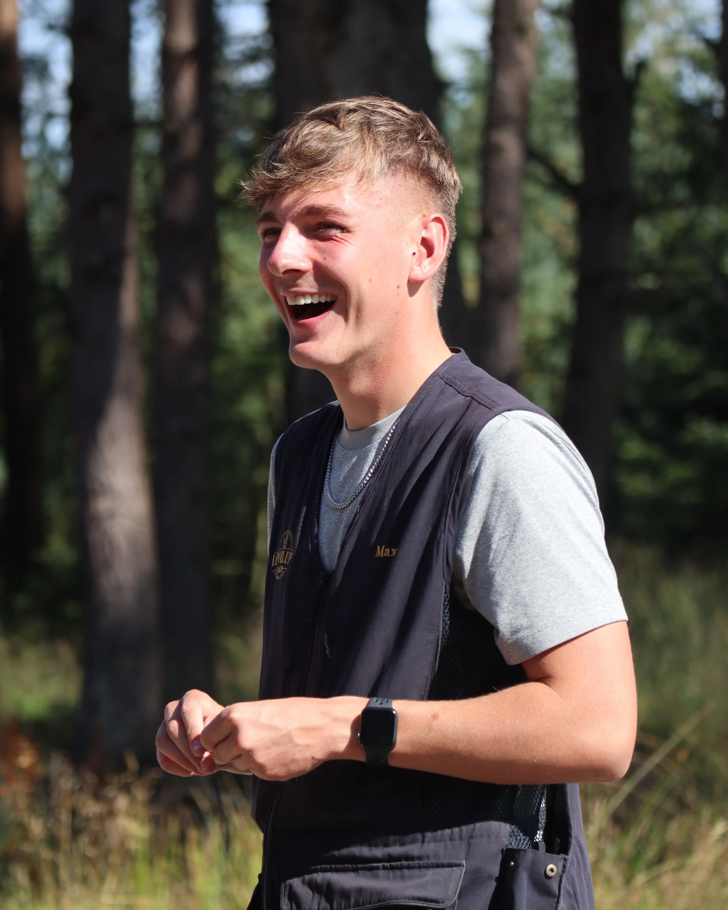 A young man with short, light brown hair smiling and looking to the side outdoors among trees, wearing a black vest over a gray T-shirt, a smartwatch on his left wrist, and a backpack.