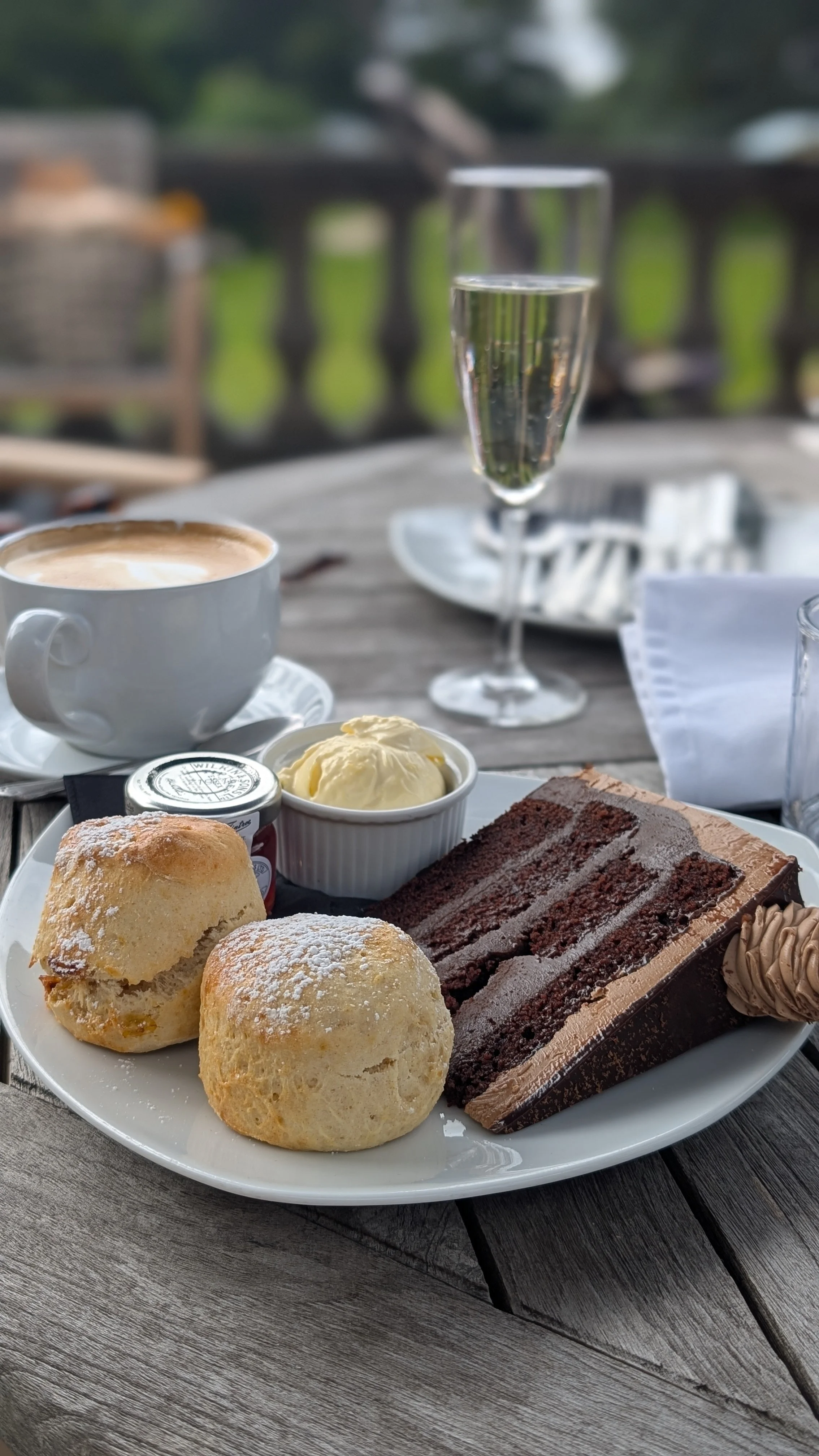 A dessert platter with two cream puffs, a slice of chocolate cake, a dollop of whipped cream, and a small jar of jam, served on a table with a cup of coffee and a glass of champagne outdoors.