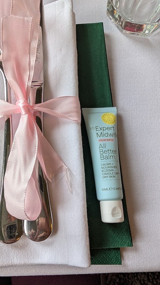 A set of utensils wrapped in a white napkin with a pink ribbon, a tube of All Better Balm, a green place card, and a glass of water on a table.