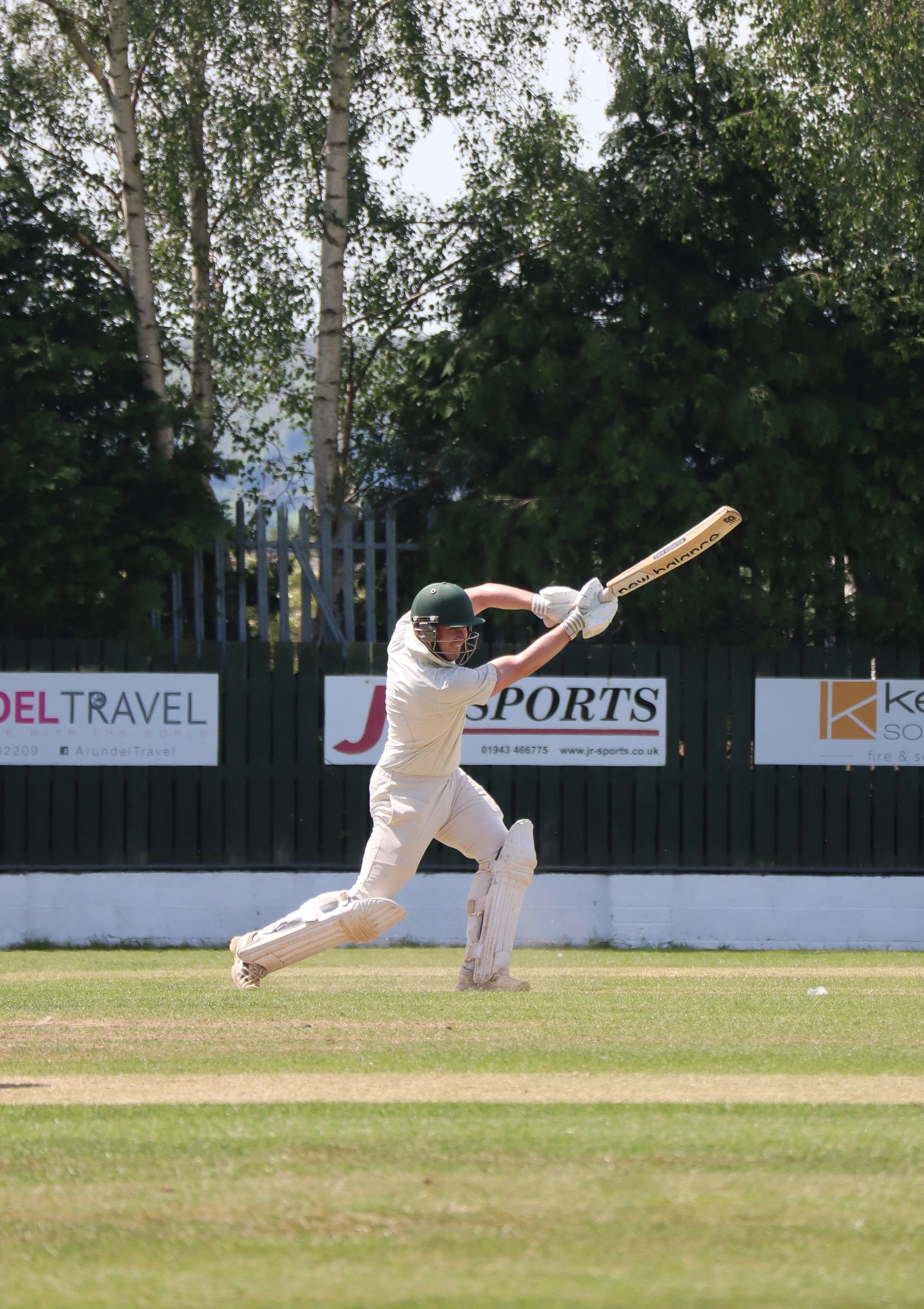 Cricket player in white uniform and green helmet hitting the ball on a cricket field with trees and advertisements in the background.