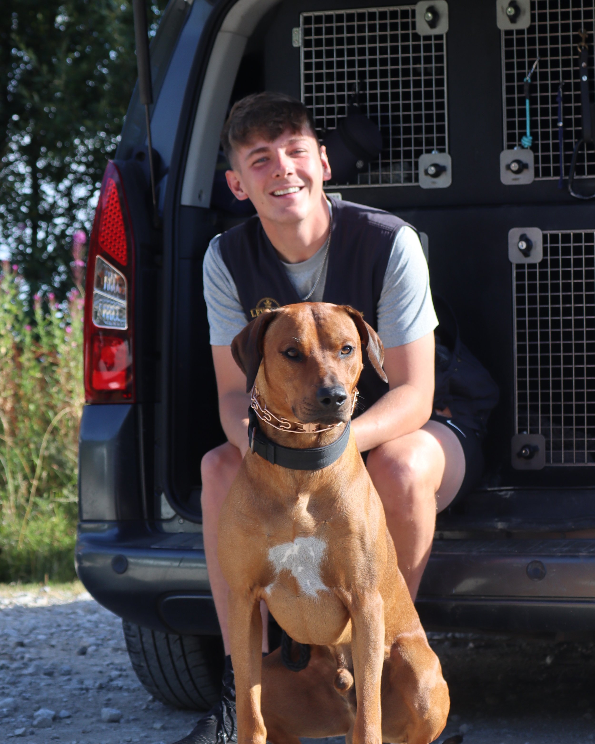A young man smiling and sitting behind a large brown dog with a black collar inside the open back of a black SUV. The dog's head and front paws are visible, and the SUV has a metal crate or divider in the back. The background includes trees and outdoor scenery.