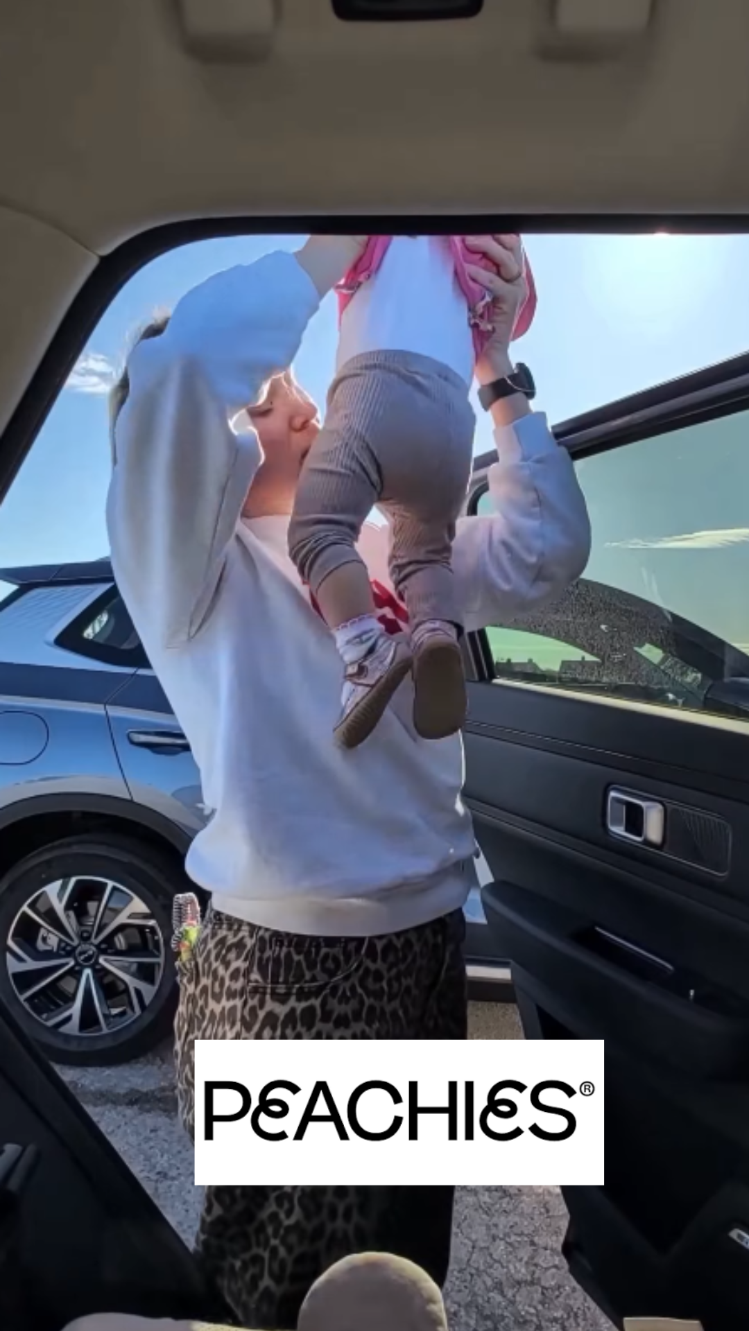 A woman wearing animal print pants and a white hoodie holding up a small child near a car window, with a blue sky in the background.
