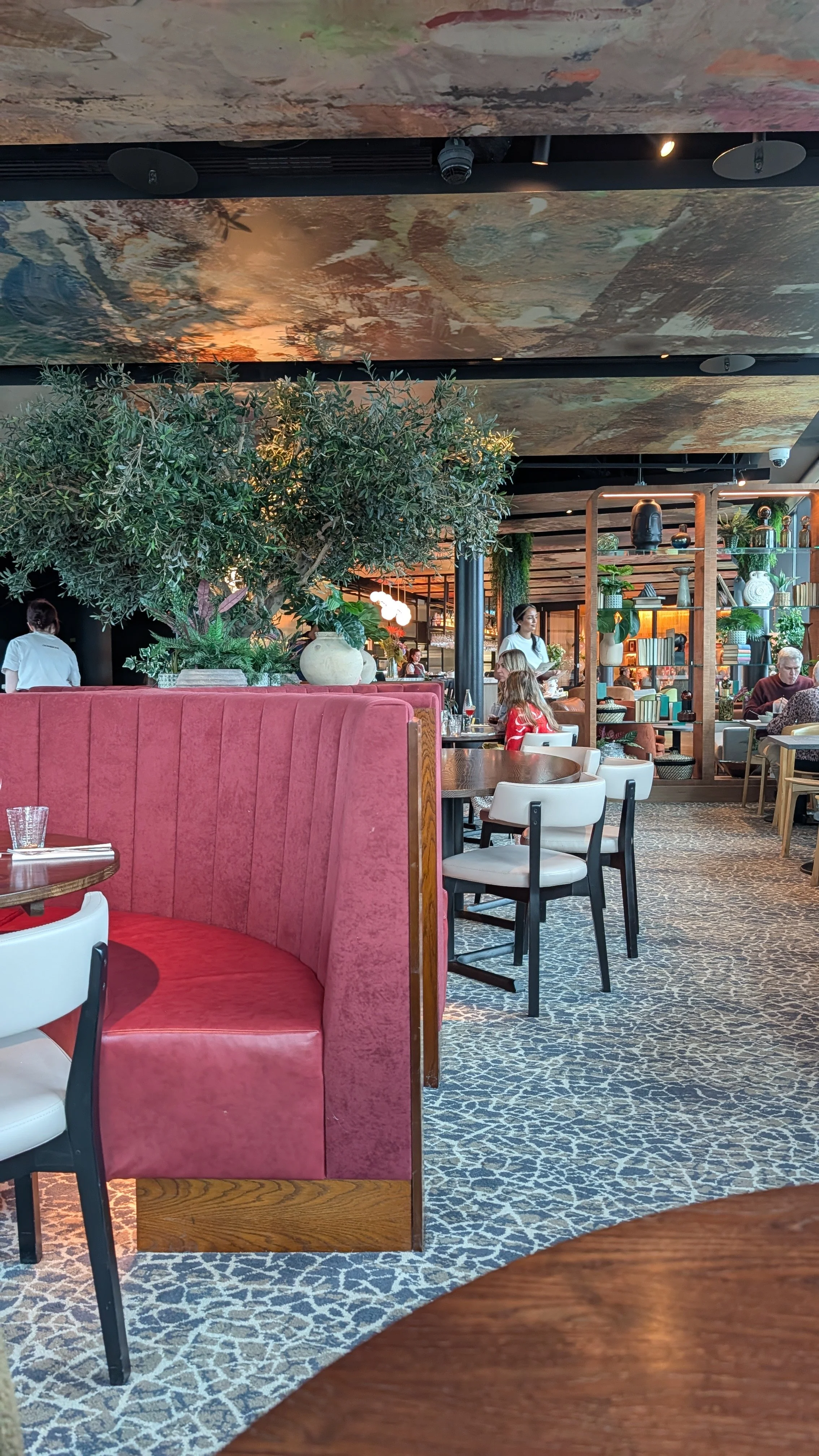 Interior view of a restaurant with various seating arrangements, plants, and decorative shelves, with people dining and staff members serving.