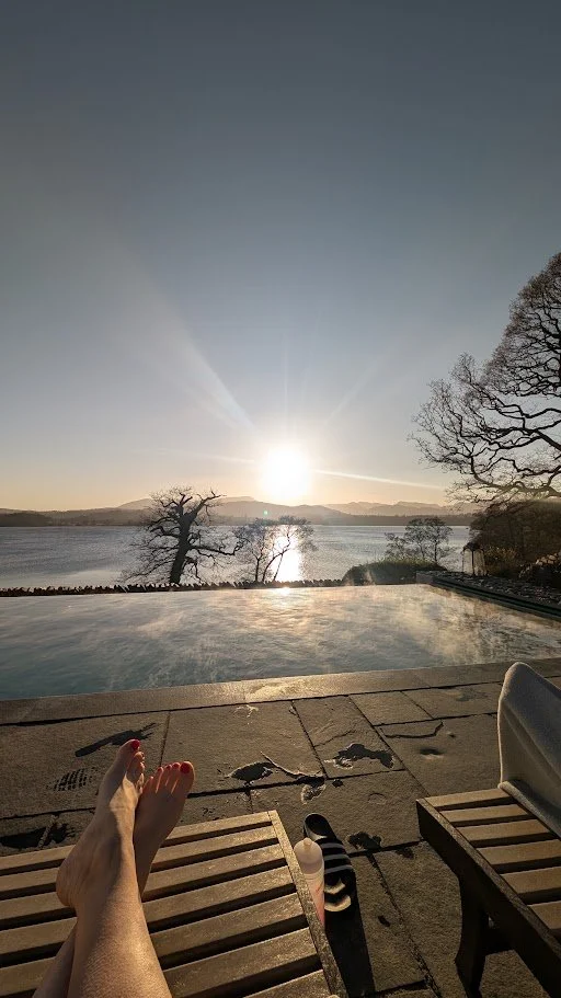 Person relaxing by a pool with legs stretched out, overlooking a lake at sunset, surrounded by trees and mountains in the distance.