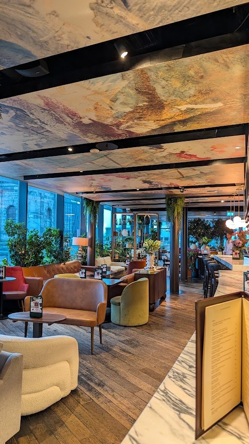 Interior of a modern restaurant or cafe with cozy seating, large windows, plants, and a marble countertop with a menu stand.