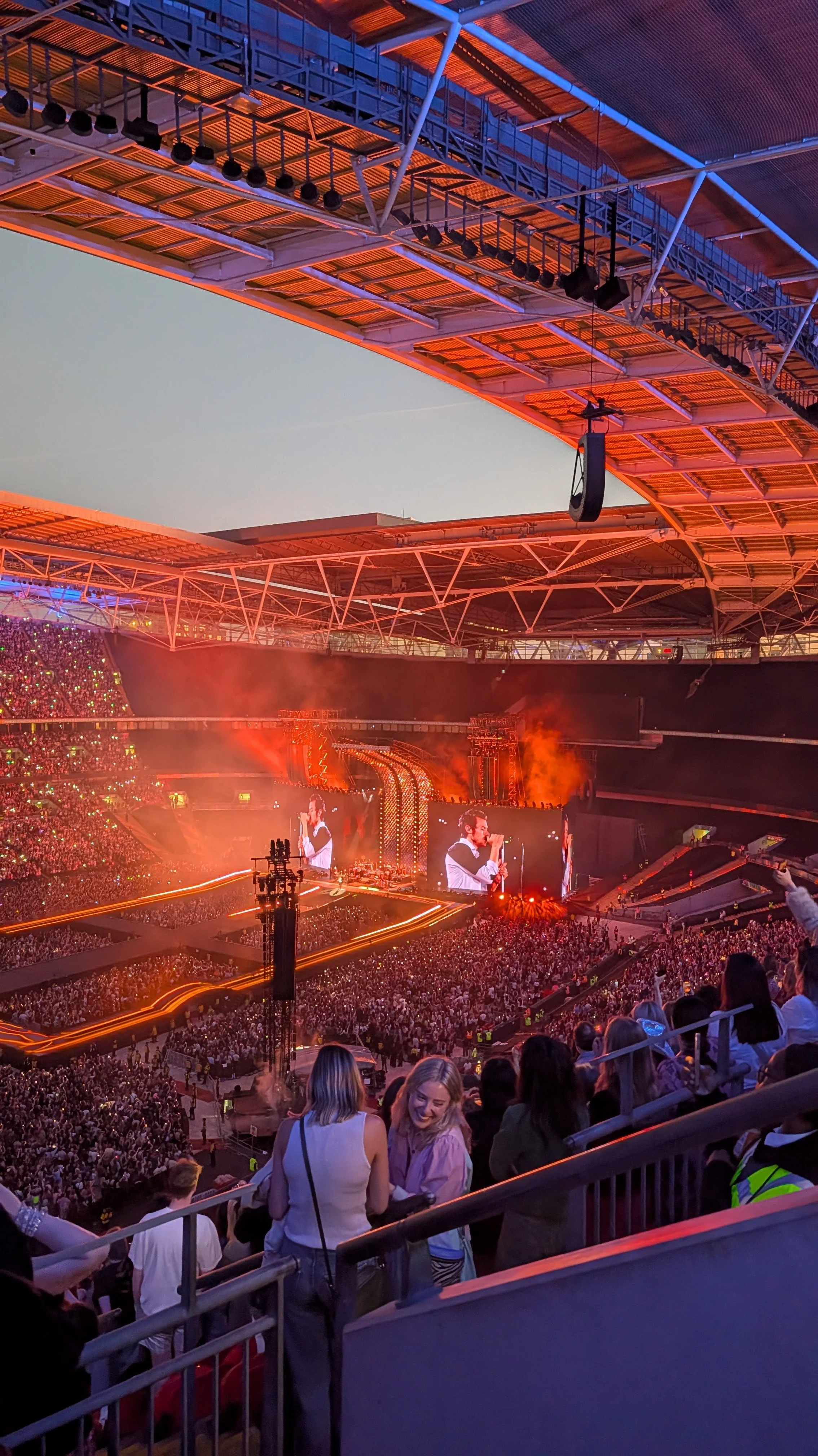Indoor stadium filled with a large audience watching a concert. The stage has bright orange lighting and large screens displaying a performer holding a microphone. The stadium structure includes a modern roof with lighting equipment hanging from it.