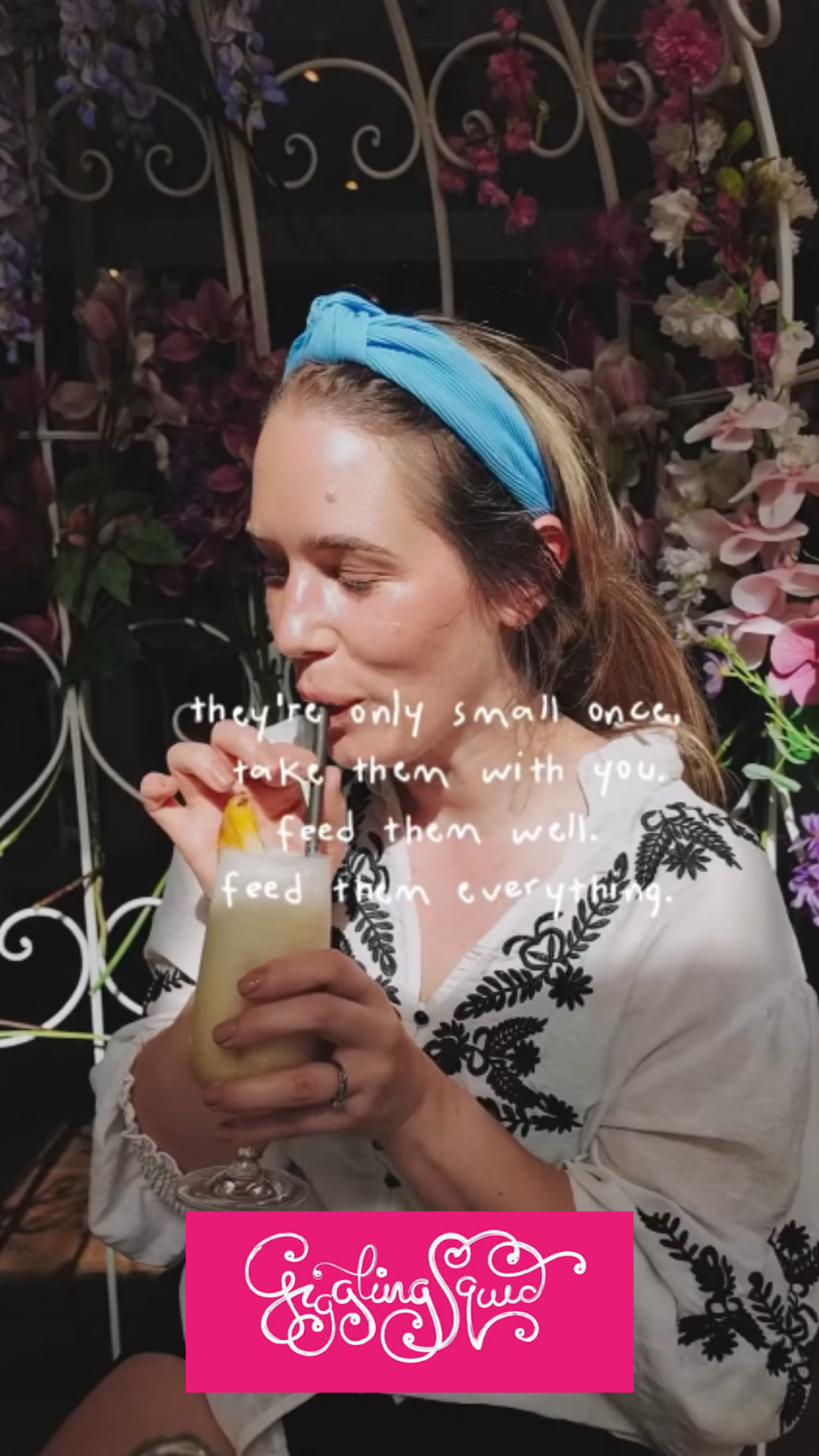 A woman with blonde hair wearing a blue headband is holding a cocktail glass with a drink and garnish, sitting in front of a floral background. Overlaid text reads, 'they're only small once, take them with you, feed them well, feed them everything.' A pink banner at the bottom says 'Cooking Guy' in white cursive.