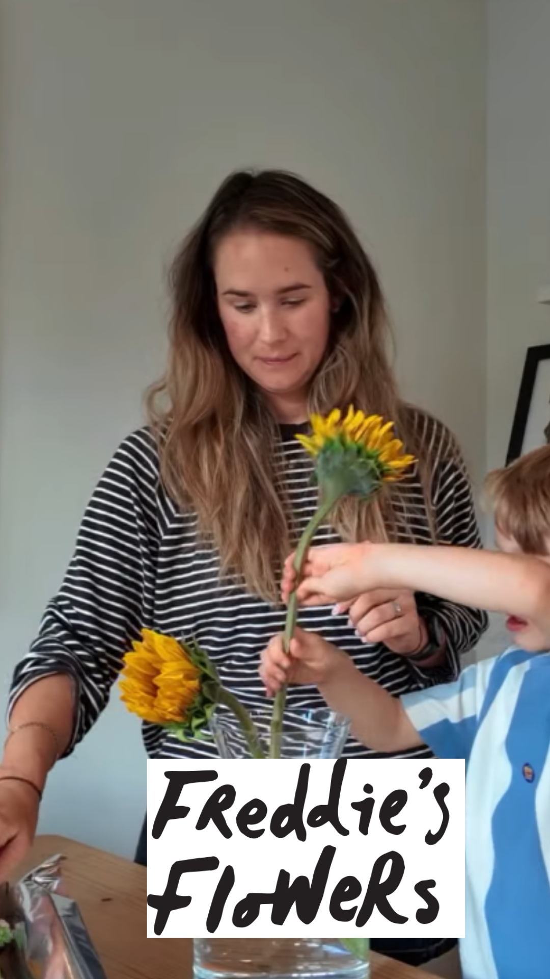 A woman with long, wavy hair receiving a sunflower from a young boy, who is reaching across a table with a sign that says "Freddie's Flowers."