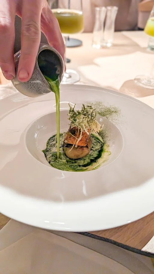Someone pouring green sauce over a gourmet dish in a white bowl at a fine dining restaurant.