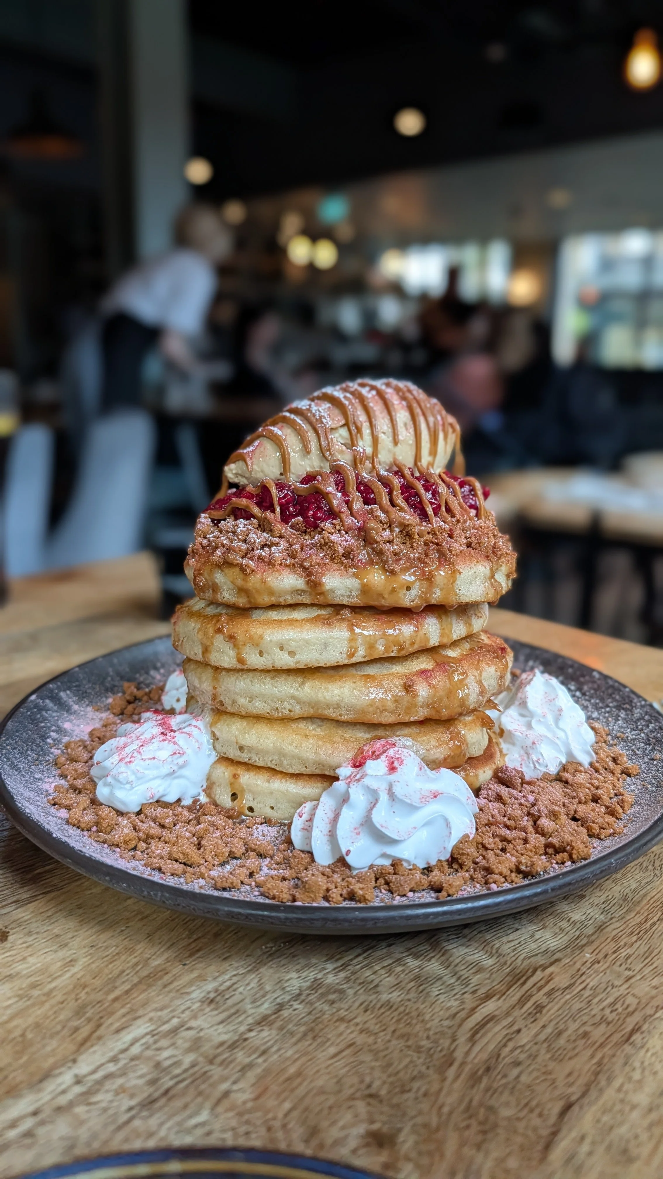 Stack of four pancakes topped with ice cream, syrup, and berries, served on a black plate with whipped cream and cookie crumbs, in a restaurant setting.