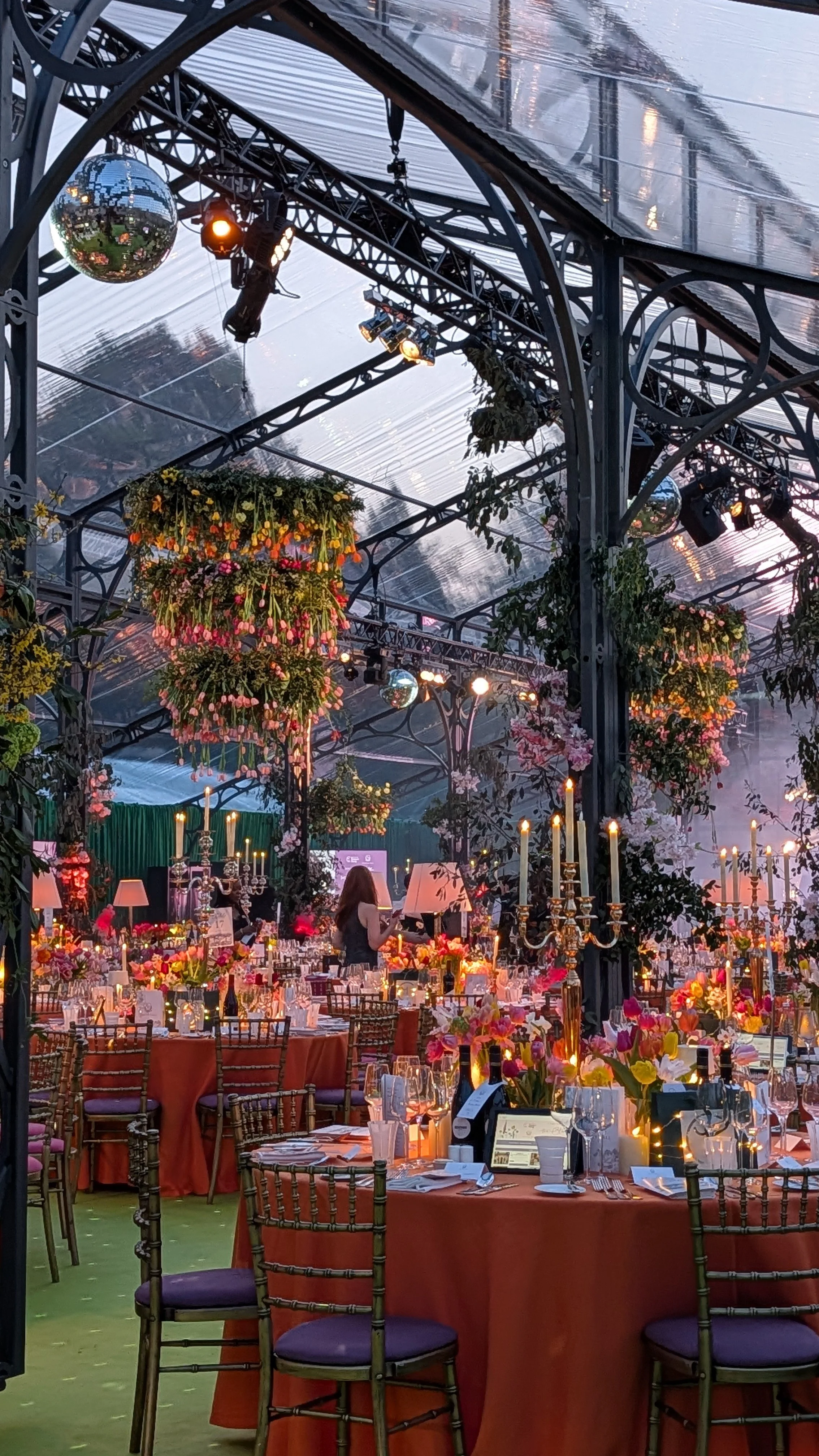 Elegant event space with decorated tables, floral arrangements, hanging greenery, chandeliers, and stage lighting inside a glass-roofed venue.